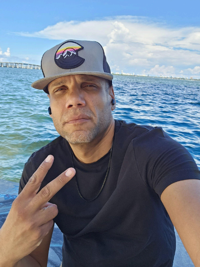 A man wearing a gray baseball cap with a mountain and rainbow logo, taking a selfie on a boat or near water, with a pier and city skyline in the background.
