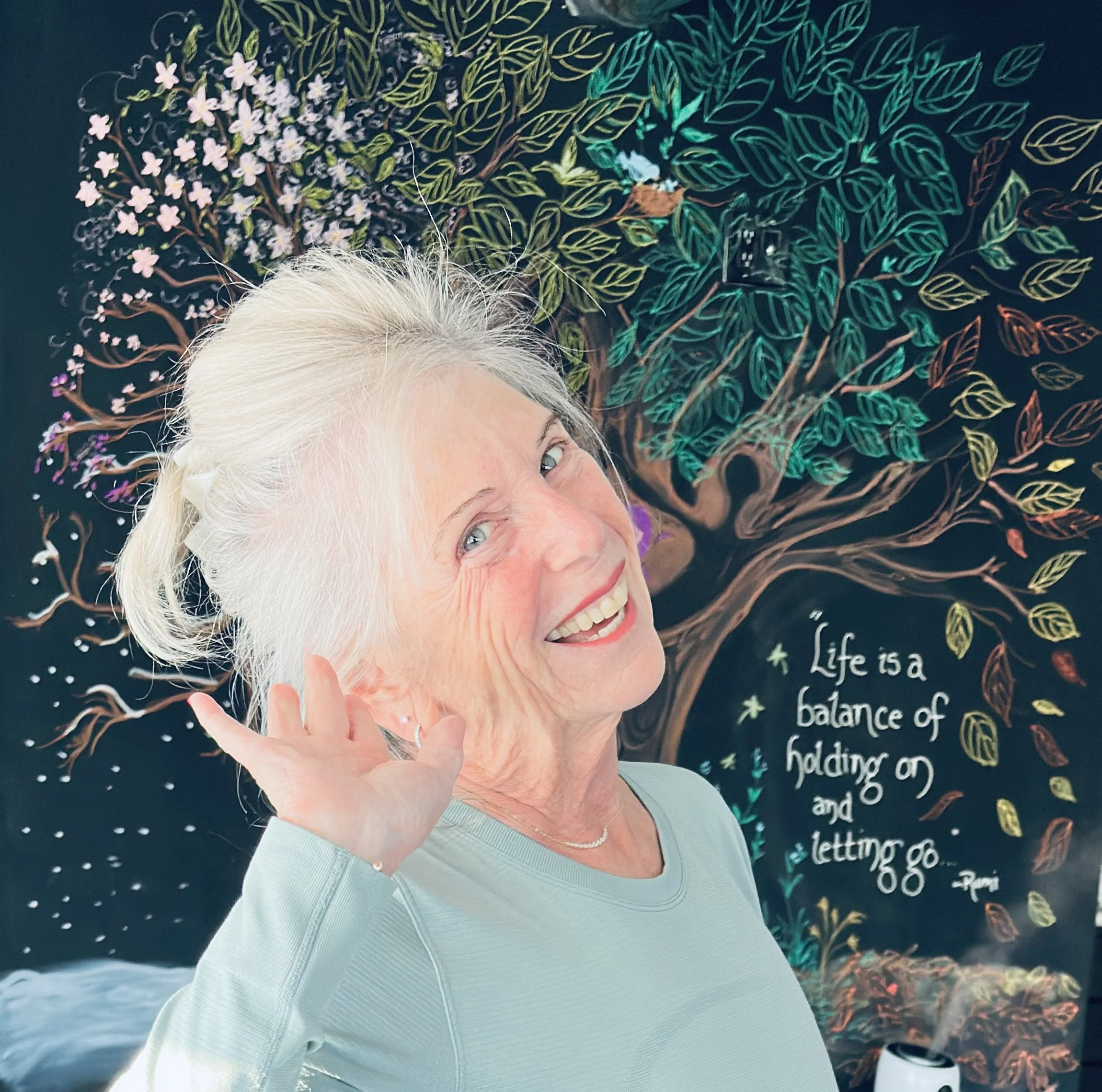 An elderly woman with white hair smiling and waving in front of a colorful chalk art tree with a quote about life.
