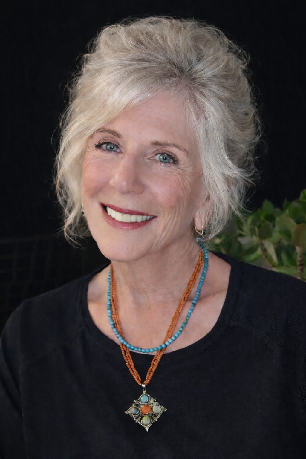 A smiling mature woman with blonde hair and blue eyes, wearing a black top and colorful beaded necklaces with a pendant, standing in front of a dark background with some green foliage.
