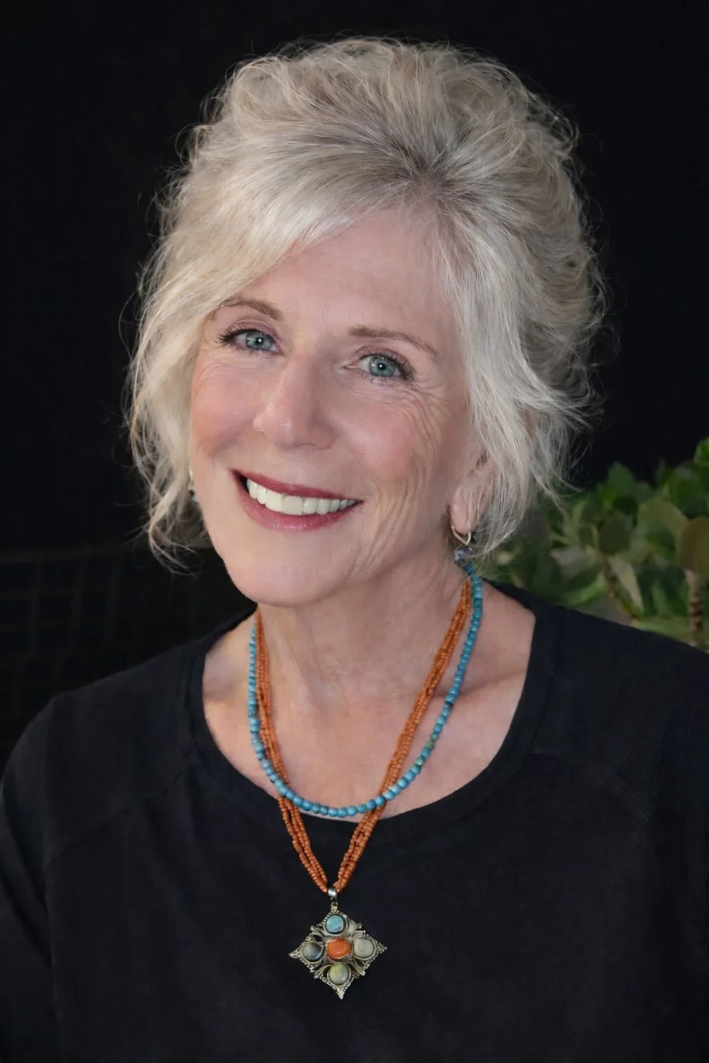 A smiling older woman with wavy, gray hair, wearing a black top and colorful beaded necklaces, standing against a dark background with some green leaves visible.