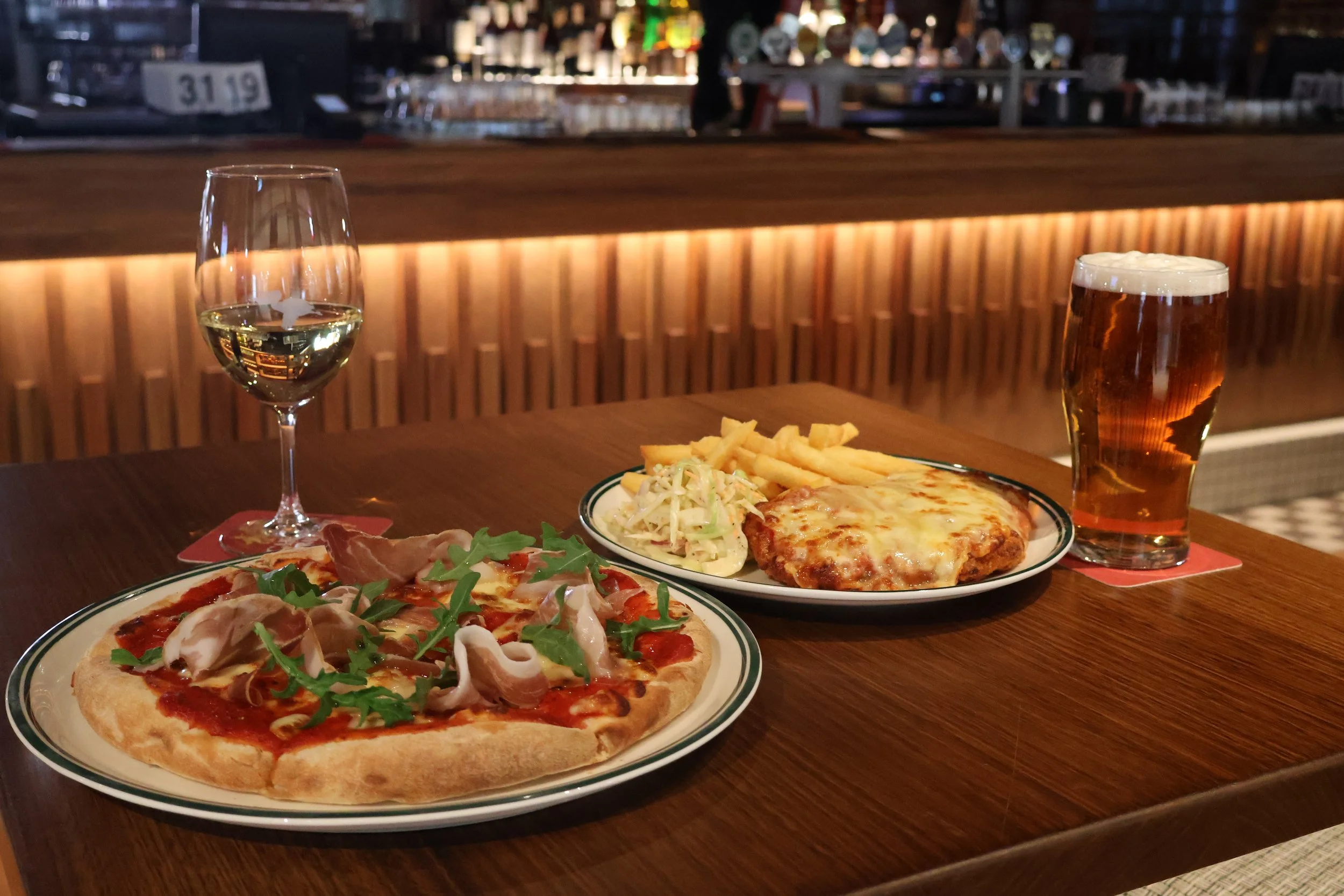 Pizza with ham, arugula, and tomato sauce, paired with a glass of white wine. Another plate with French fries, coleslaw, a cheeseburger patty, and a pint of beer. The background is a bar with drinks.