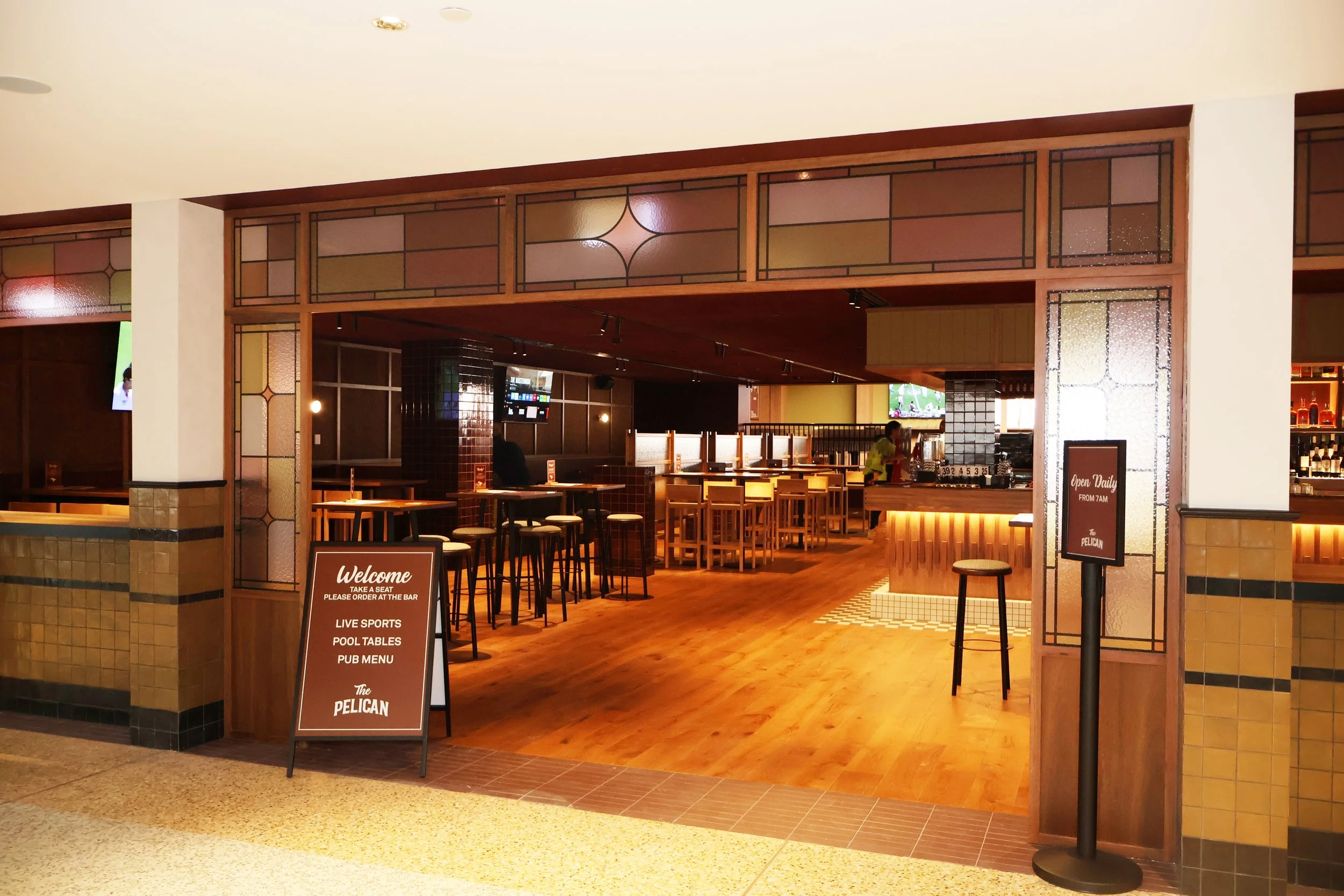 Interior of a sports bar called The Pelican, with a welcoming sign, bar counter, high tables, chairs, and TVs showing sports, featuring warm lighting and wooden decor.