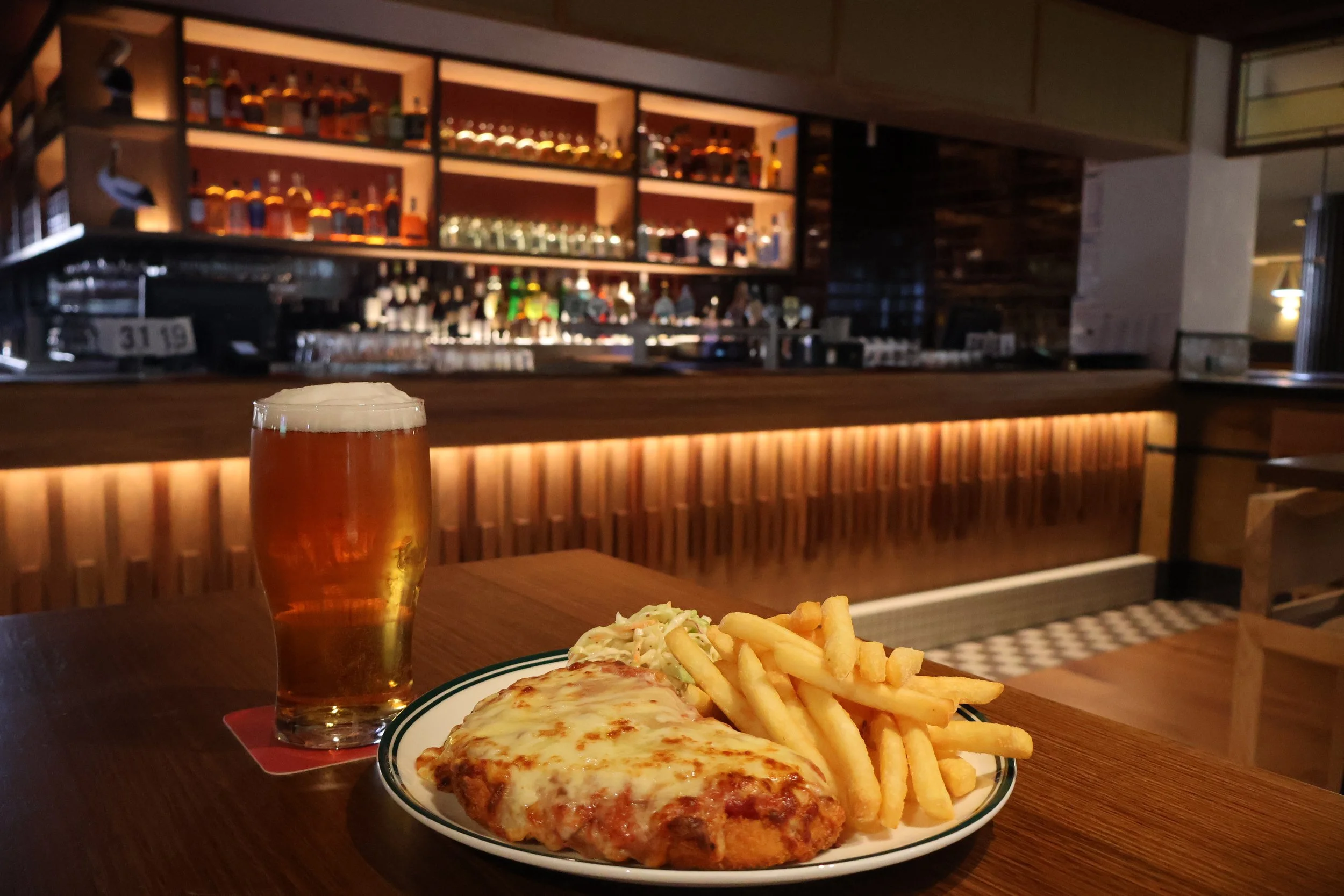 A plate of cheese pizza, French fries, and coleslaw on a wooden table with a glass of beer in a dimly lit bar or restaurant.