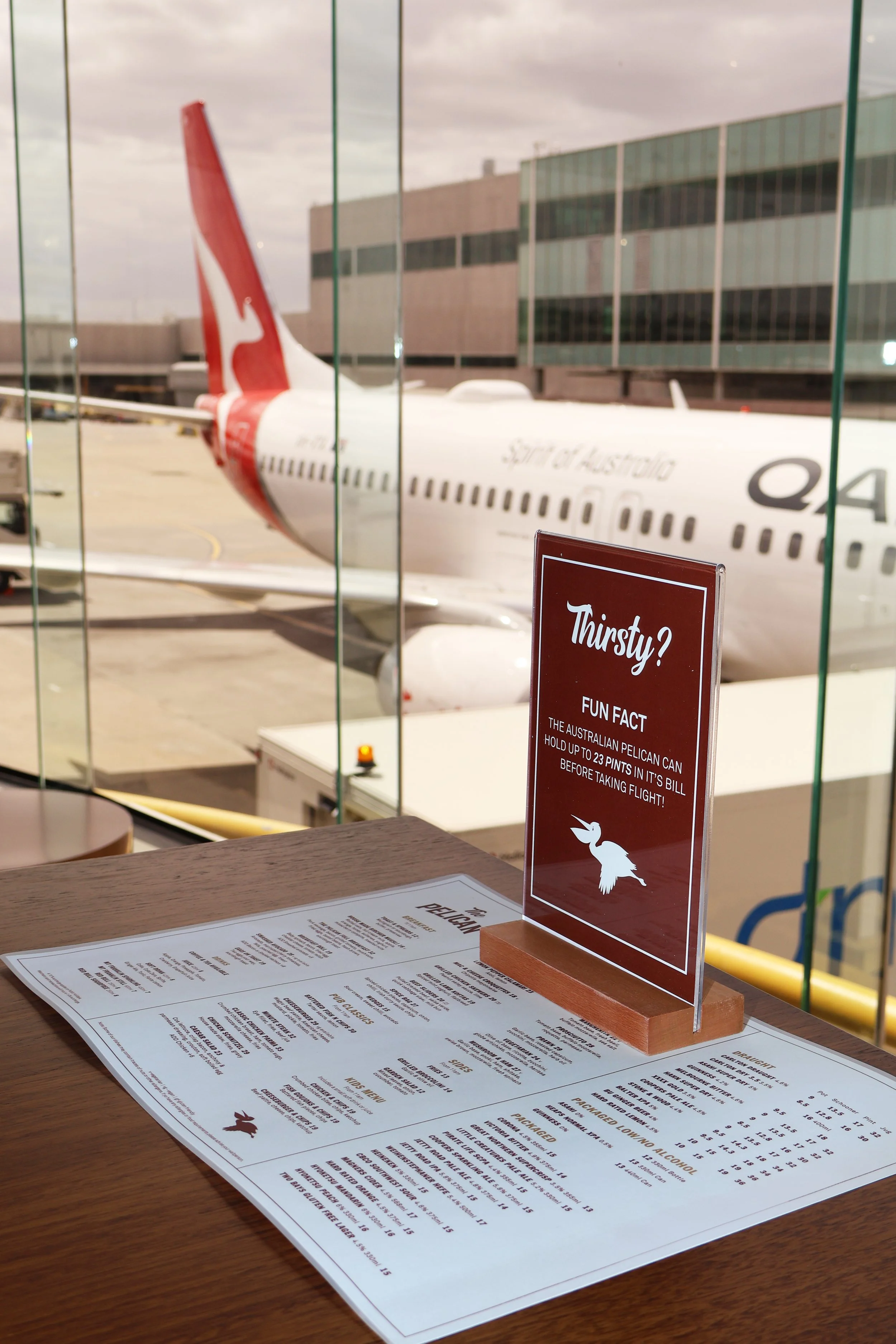 View of an airplane parked at the gate from inside a windowed cafe. A menu and a sign that says "Thirsty? Fun Fact: The Australian pelican can hold up to 23 pints in its bill before taking flight!" are on the table.