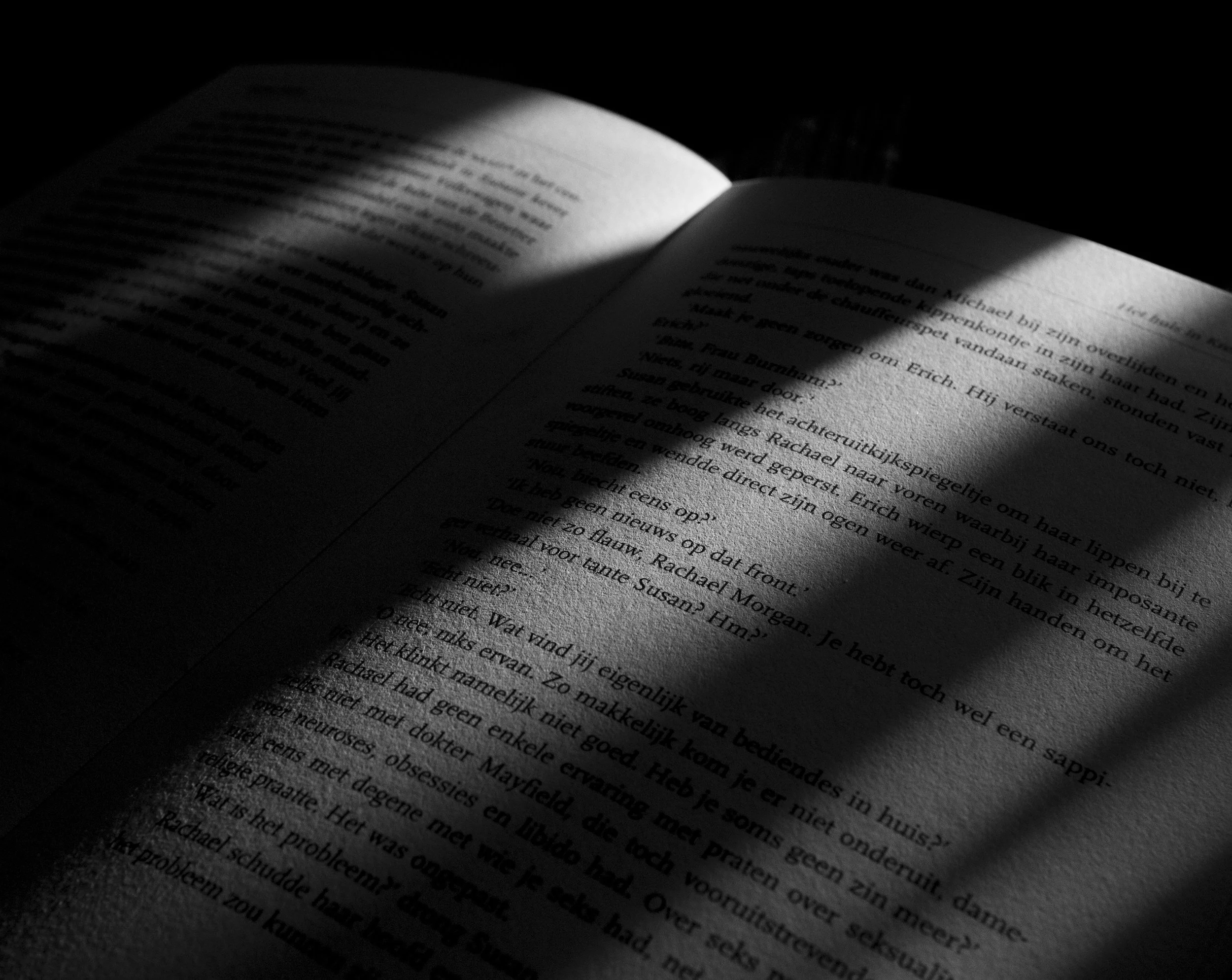 Open book with text in Dutch, illuminated by a narrow beam of light, creating a shadow on the left side and highlighting the right side of the pages.