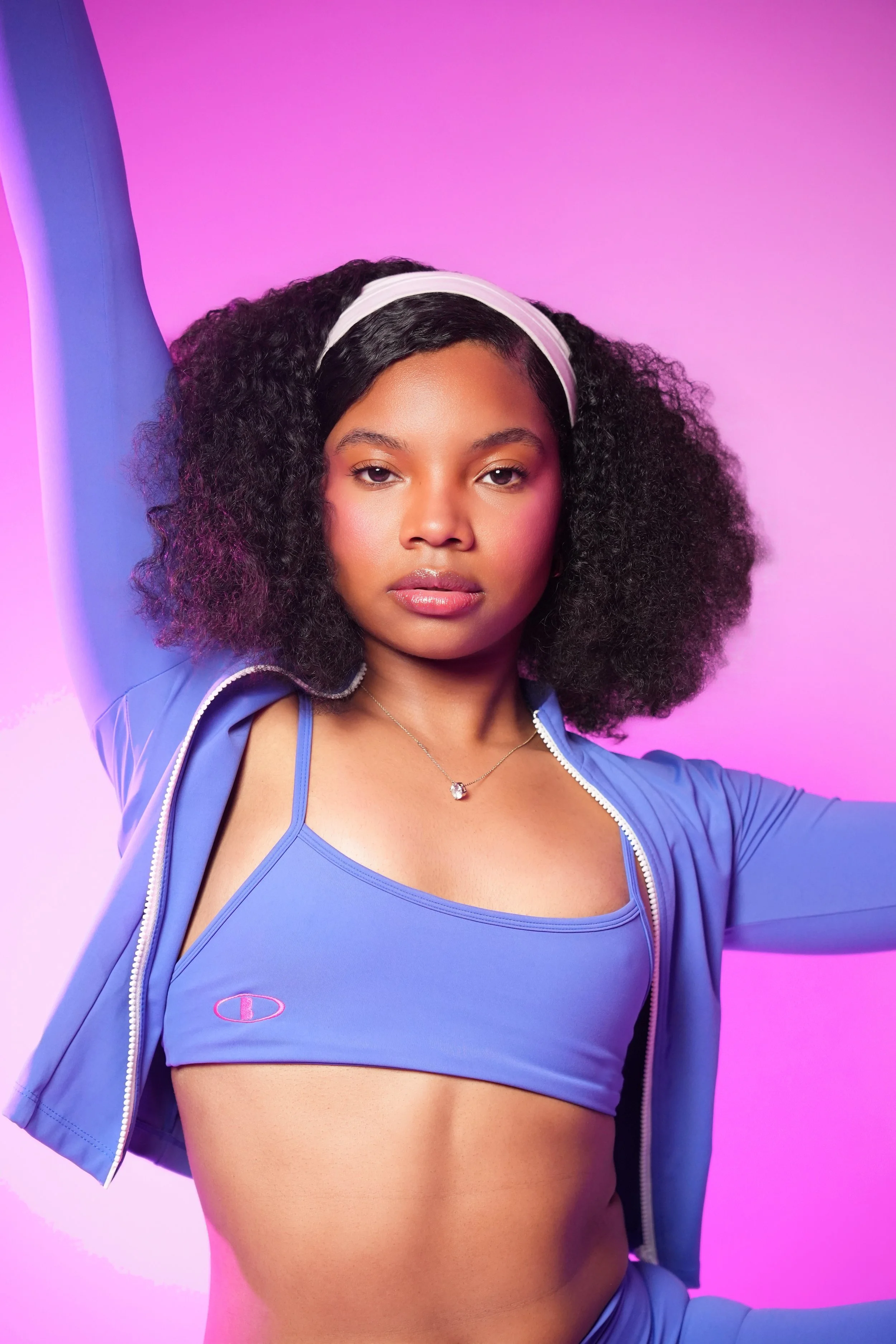 A young woman with curly hair wearing a blue sports bra and jacket, posing with one arm raised against a pink and purple background.