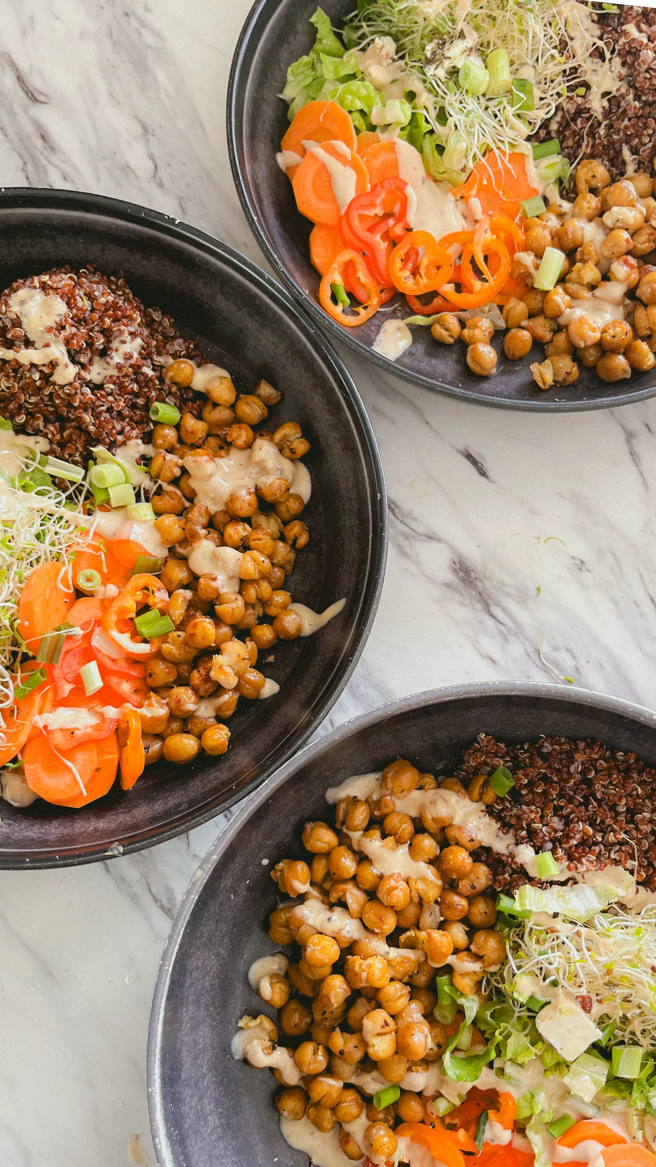 Three black bowls containing salad with chickpeas, red quinoa, cherry tomatoes, shredded lettuce, green onions, sprouts, and a creamy dressing, on a white marble surface.