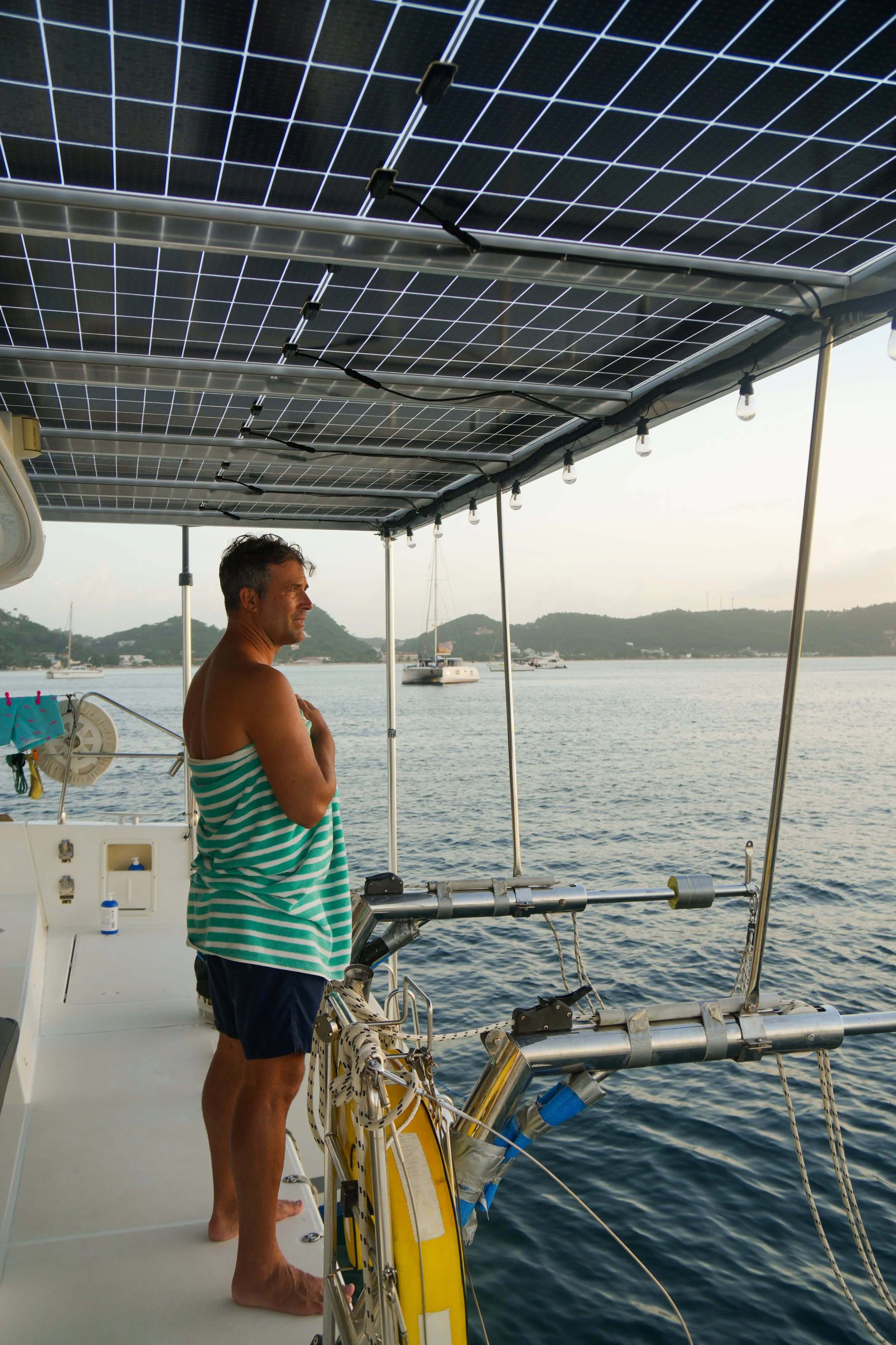 A man standing on a boat deck underneath large solar panels, looking out over the water with boats and islands in the background.