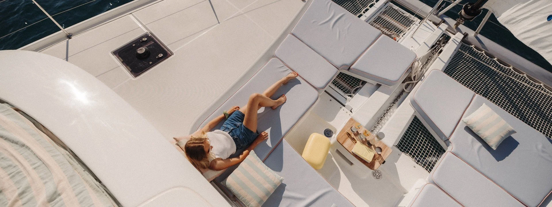 An overhead view of a woman relaxing on a cushioned lounge on a yacht with a small table holding drinks, against the backdrop of the ocean.