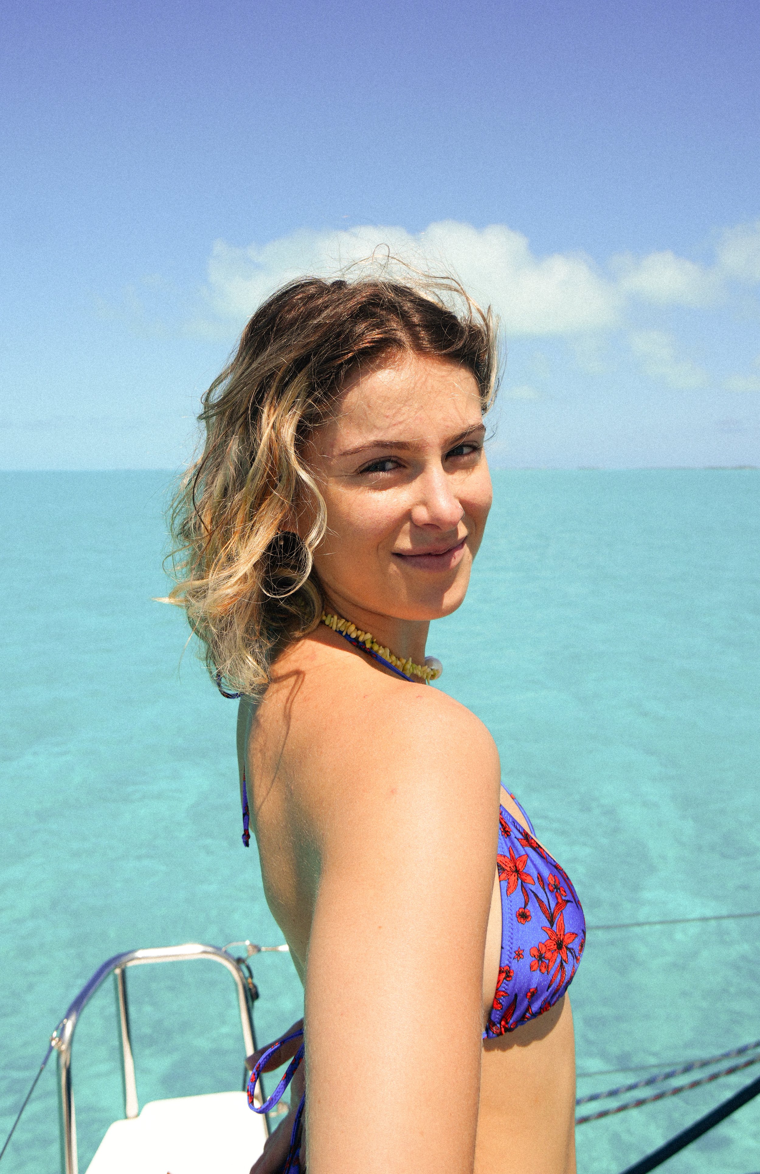 A woman in a floral blue bikini top smiling on a boat with turquoise water and blue sky in the background.