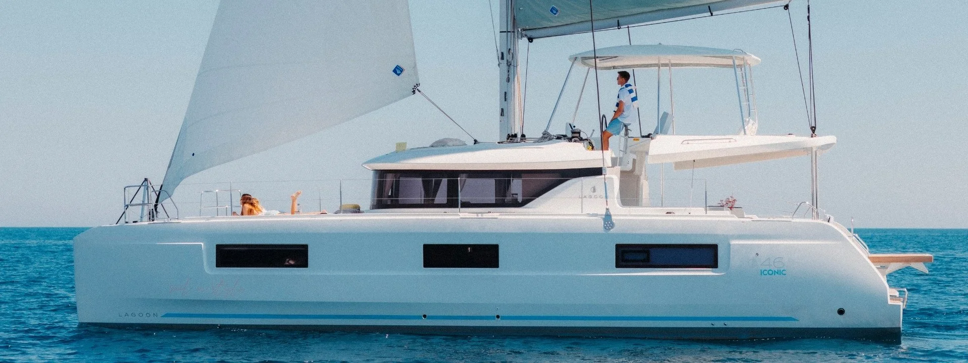 A white luxury sailboat on the open water with a man standing on the upper deck and a woman lying on the sunbathing area, both enjoying a sunny day.