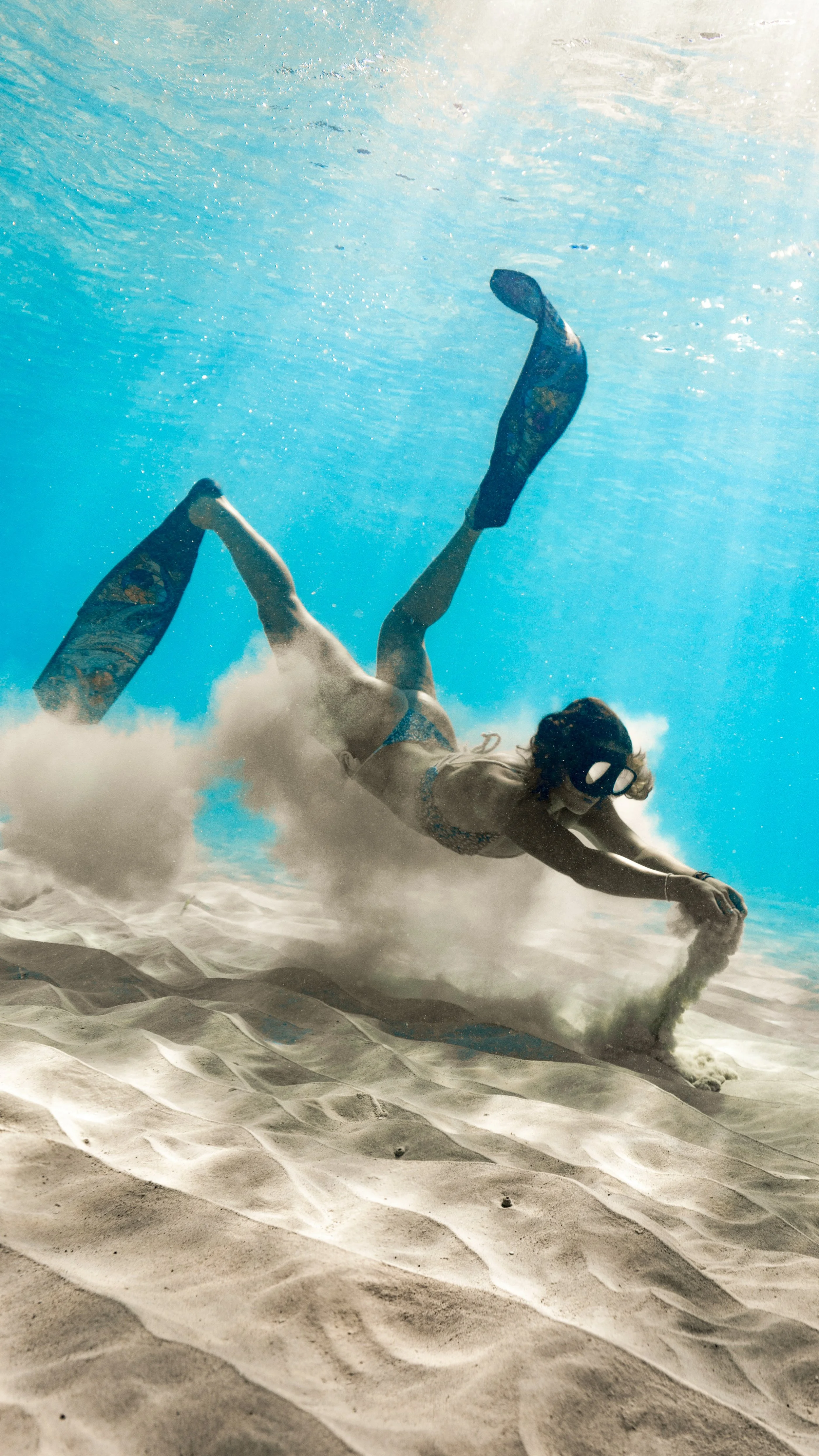 A woman in a bikini with colorful leggings and goggles is diving into the water, creating a splash on a sandy bottom in a swimming pool.