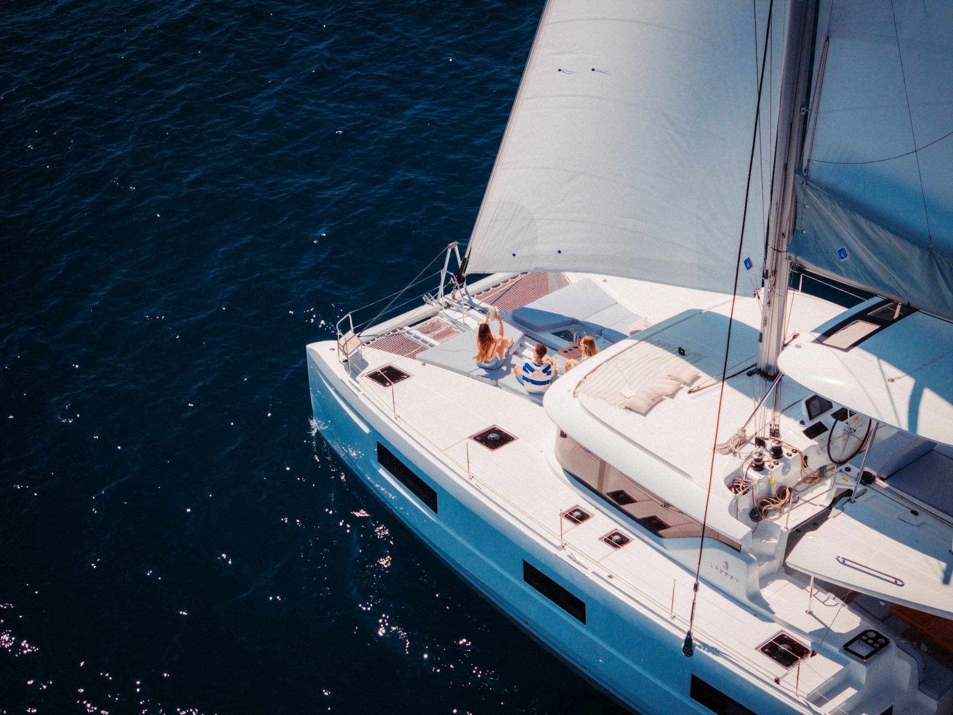 Aerial view of a white yacht sailing on dark blue water with three people relaxing on the deck.