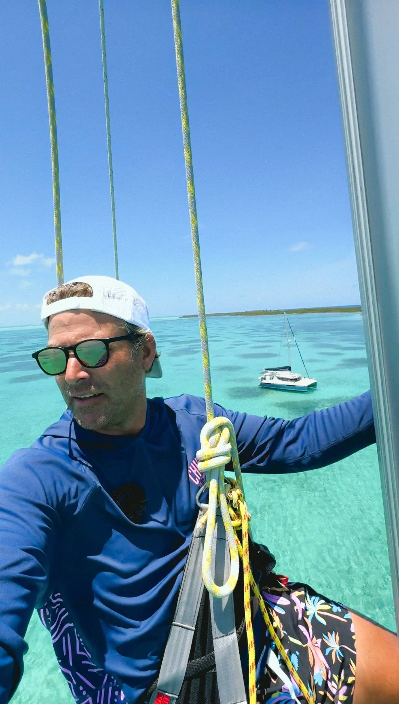 Man taking a selfie on a boat in turquoise waters, wearing sunglasses, a white cap backwards, a blue long-sleeve shirt, and colorful shorts, with sailing ropes and a sailboat in the background.