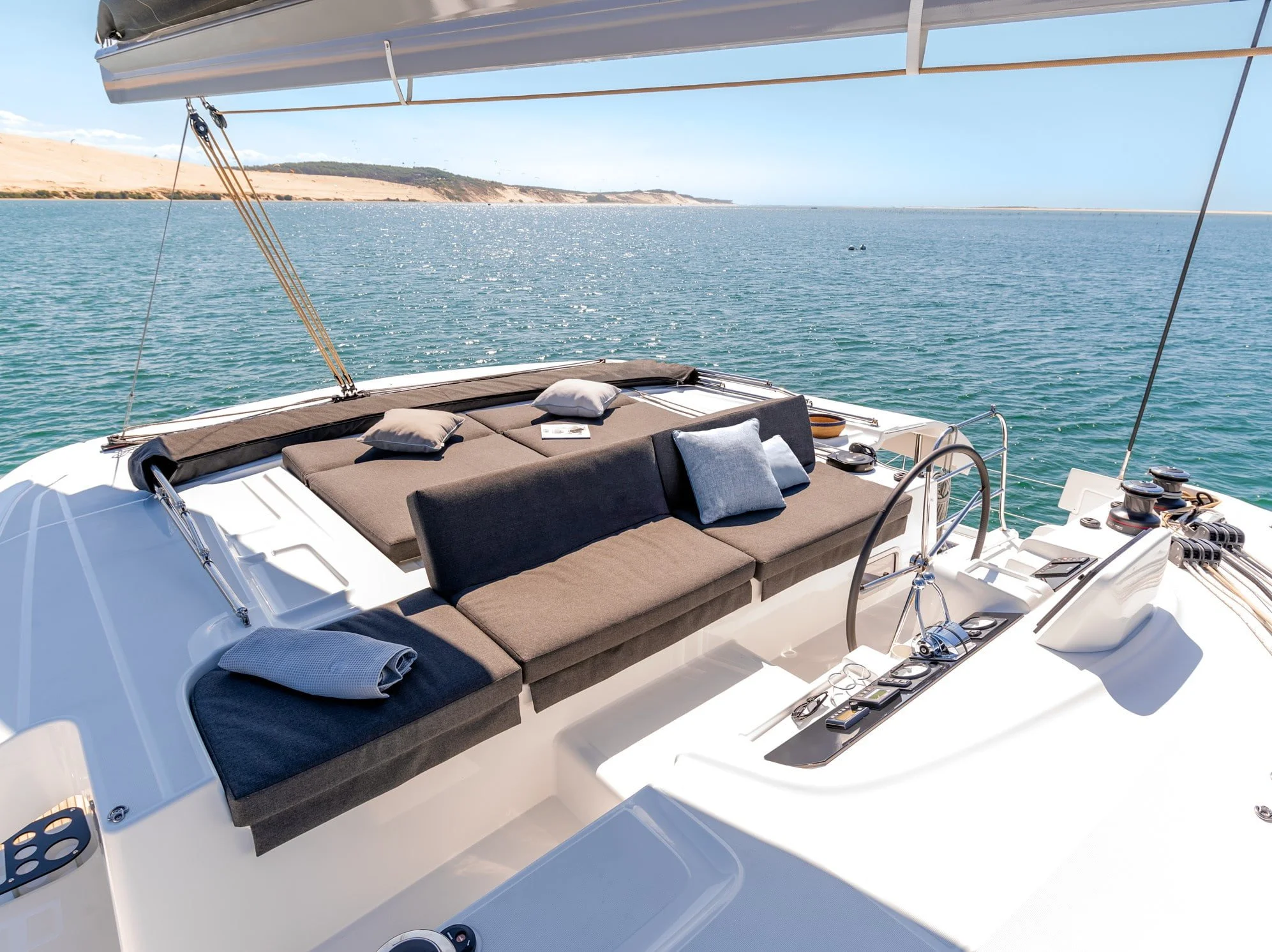 The deck of a luxurious white yacht with cushioned seating, pillows, and a view of the ocean and distant land.