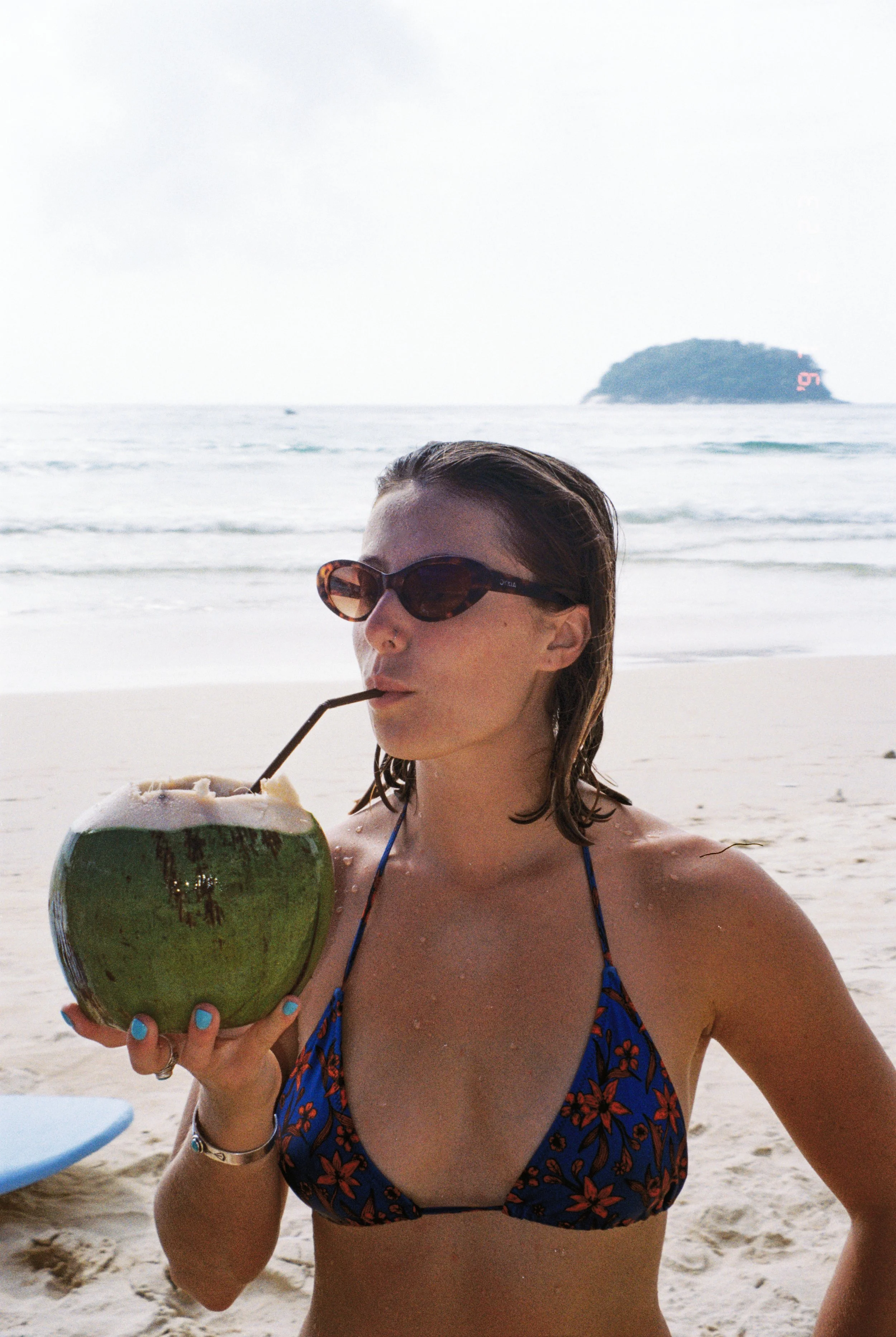 A woman in a blue floral bikini and sunglasses drinks from a coconut with a straw on a beach with ocean waves and an island in the background.