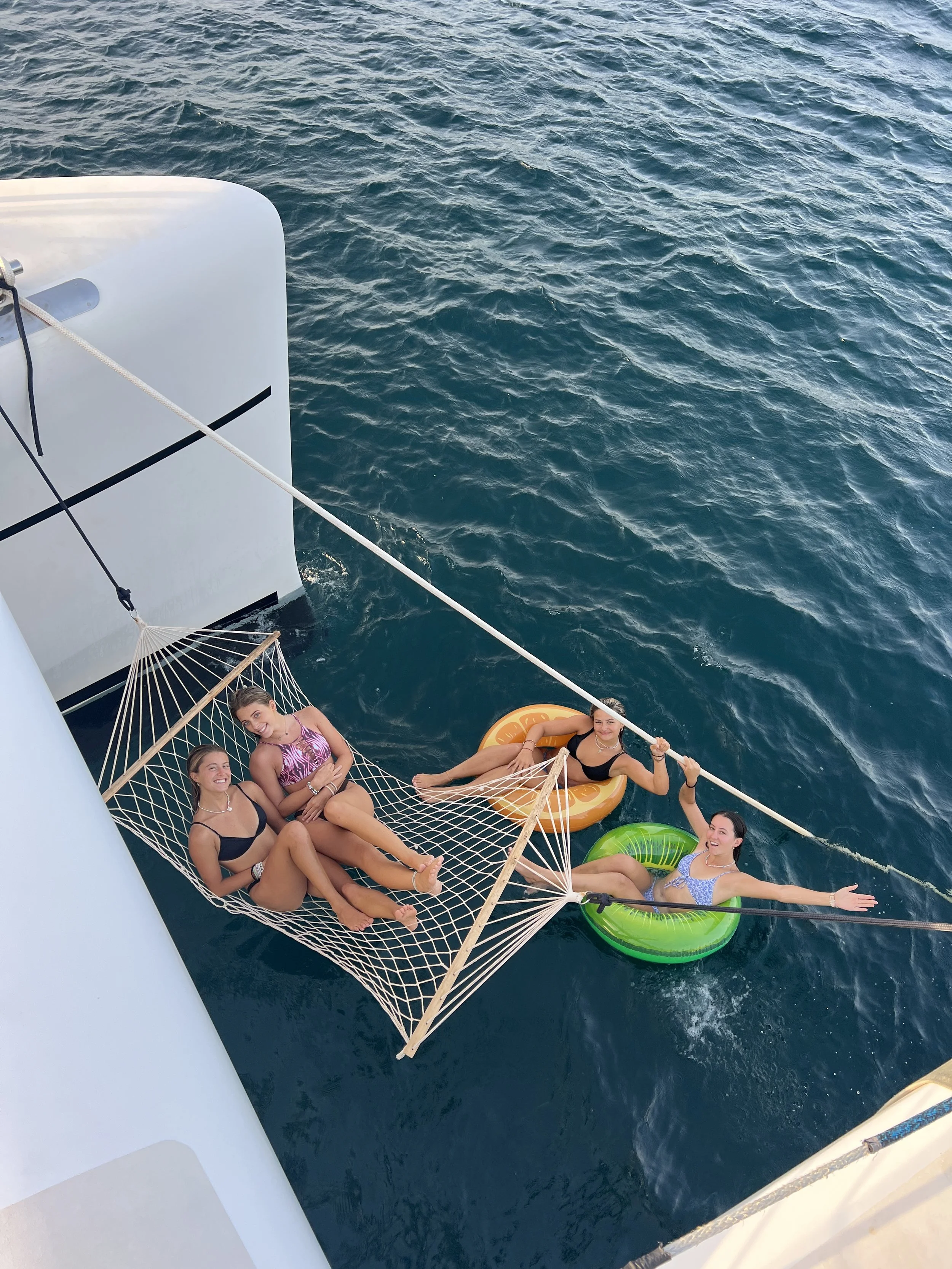 Four women relaxing on a hammock and in pool floats hanging off a boat in the ocean.