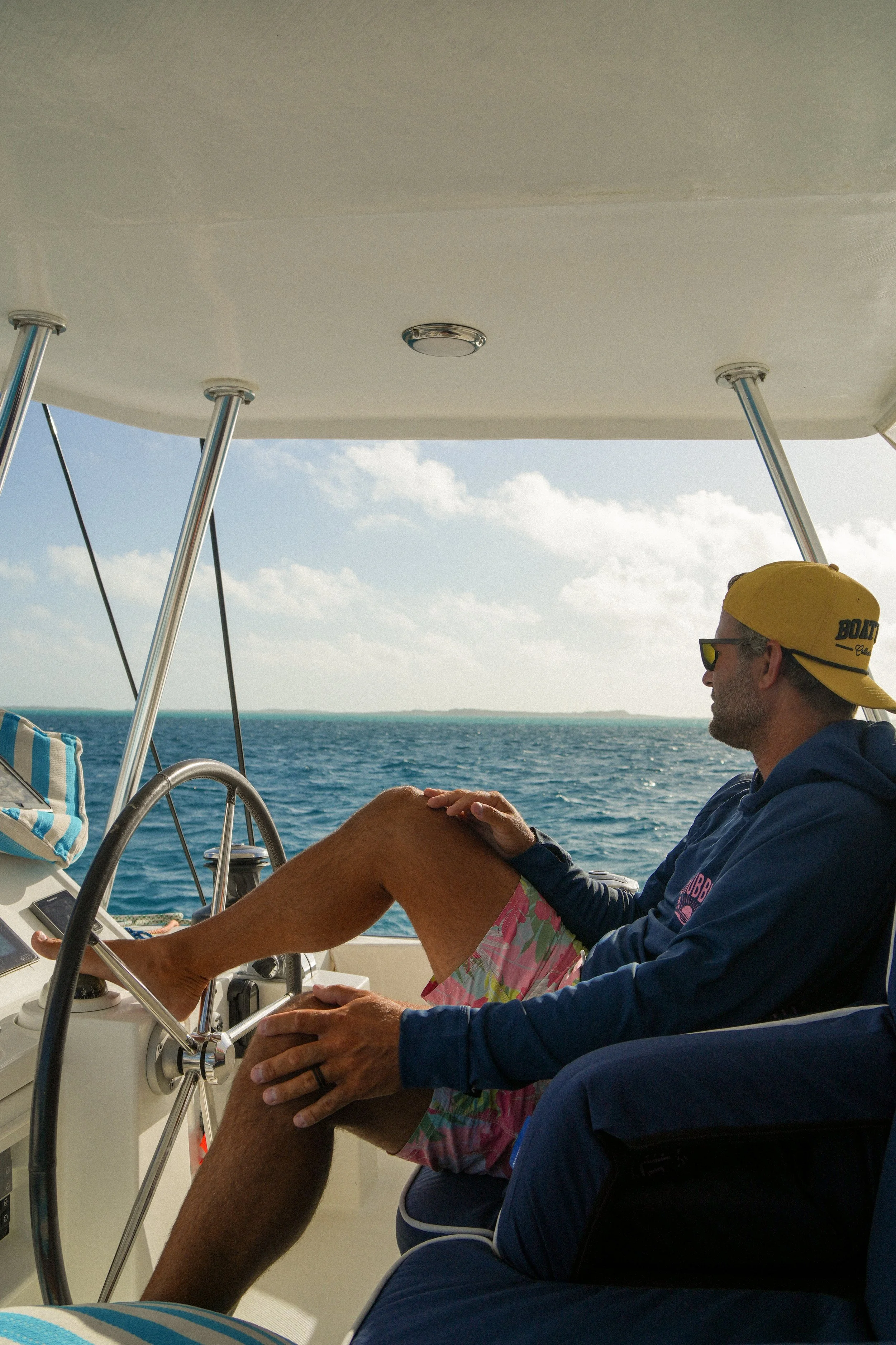 A man relaxing on a boat, sitting in a chair with one leg raised, wearing colorful shorts, a dark hoodie, sunglasses, and a yellow cap backwards. The boat is on the water under a partly cloudy sky.