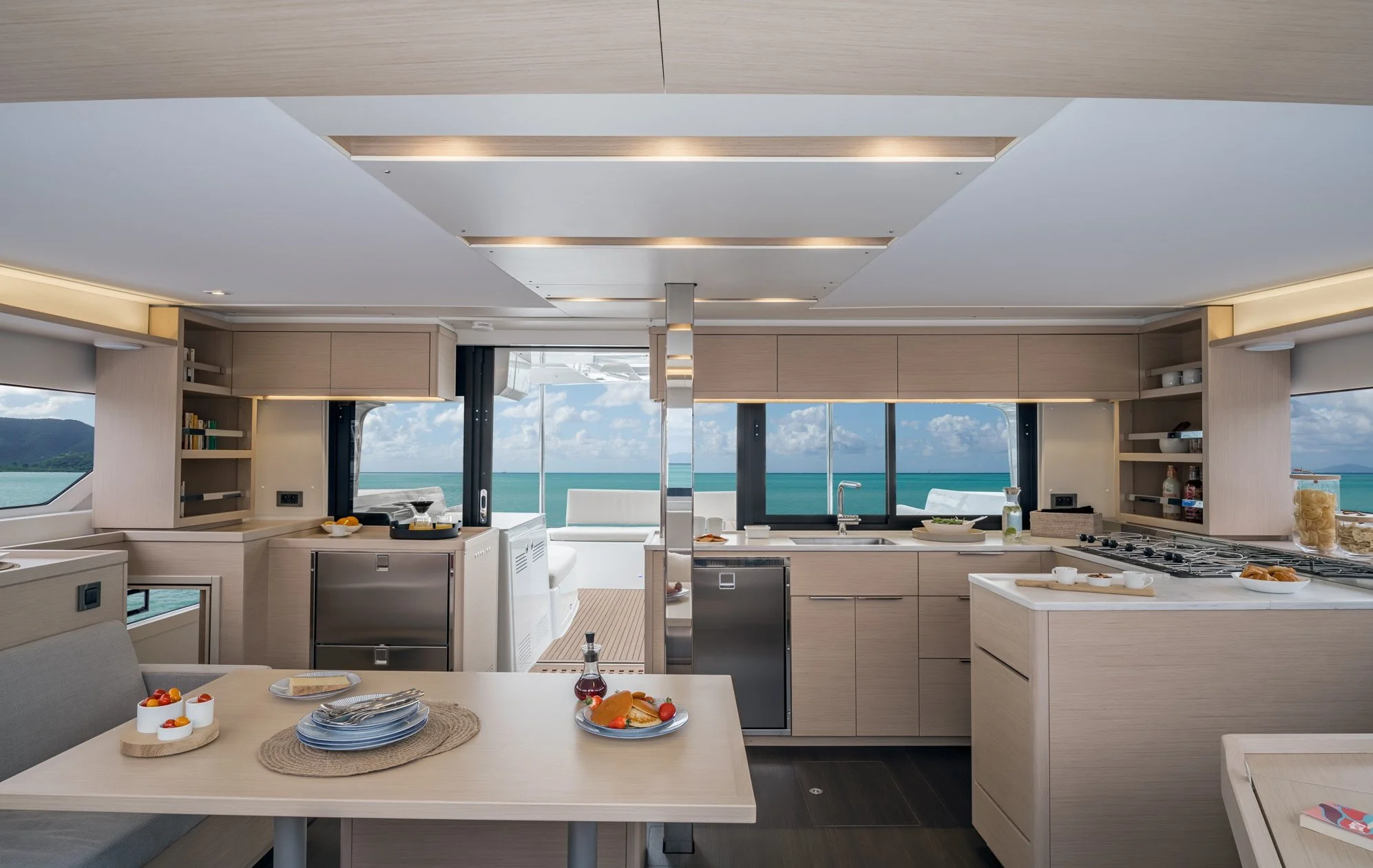 Modern kitchen with large windows overlooking the ocean, featuring light wood cabinets, a white countertop, and a dining table with dishes and glasses, with a view of the sea and blue sky.