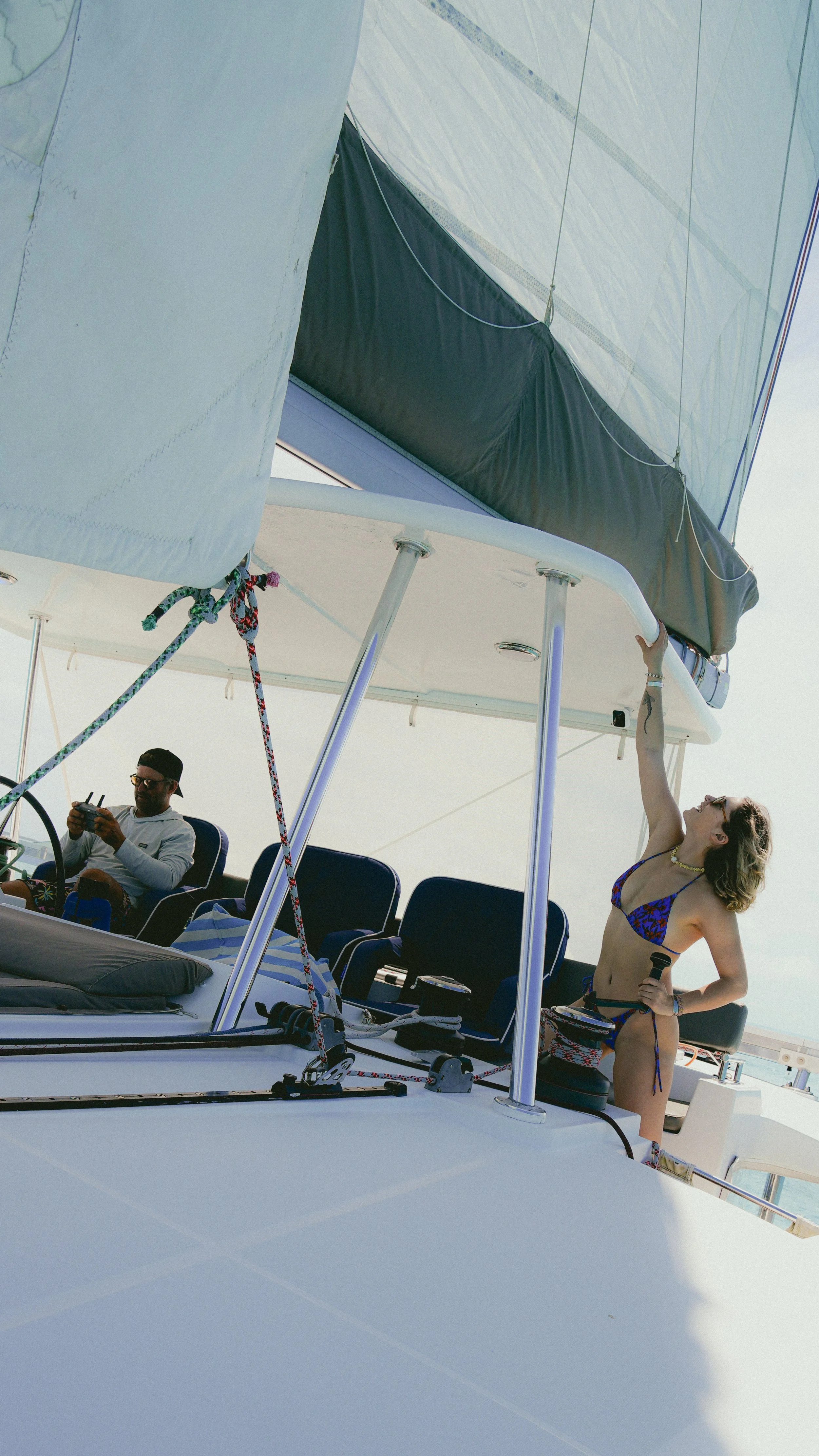 A woman in a bikini on a sailboat, standing and holding onto the sail's edge while a man is sitting wearing sunglasses and a cap, operating a remote control, on a bright day.