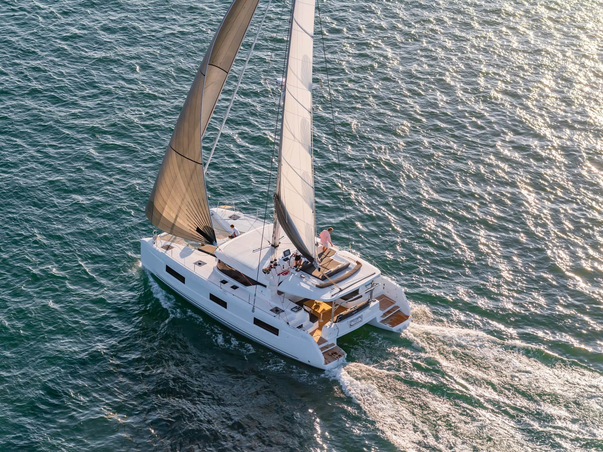 An aerial view of a white sailing yacht with brown sails, cruising on the ocean during sunset.