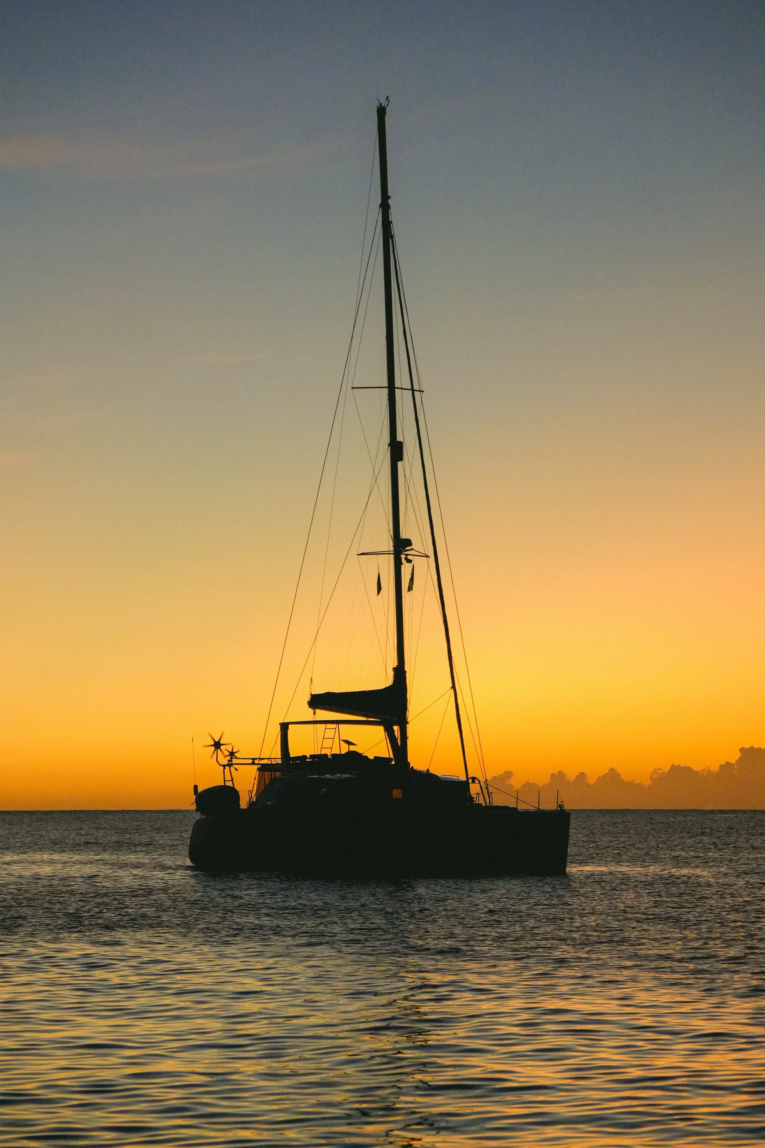 Silhouette of a sailboat on the water at sunset with a colorful sky and small clouds in the background.