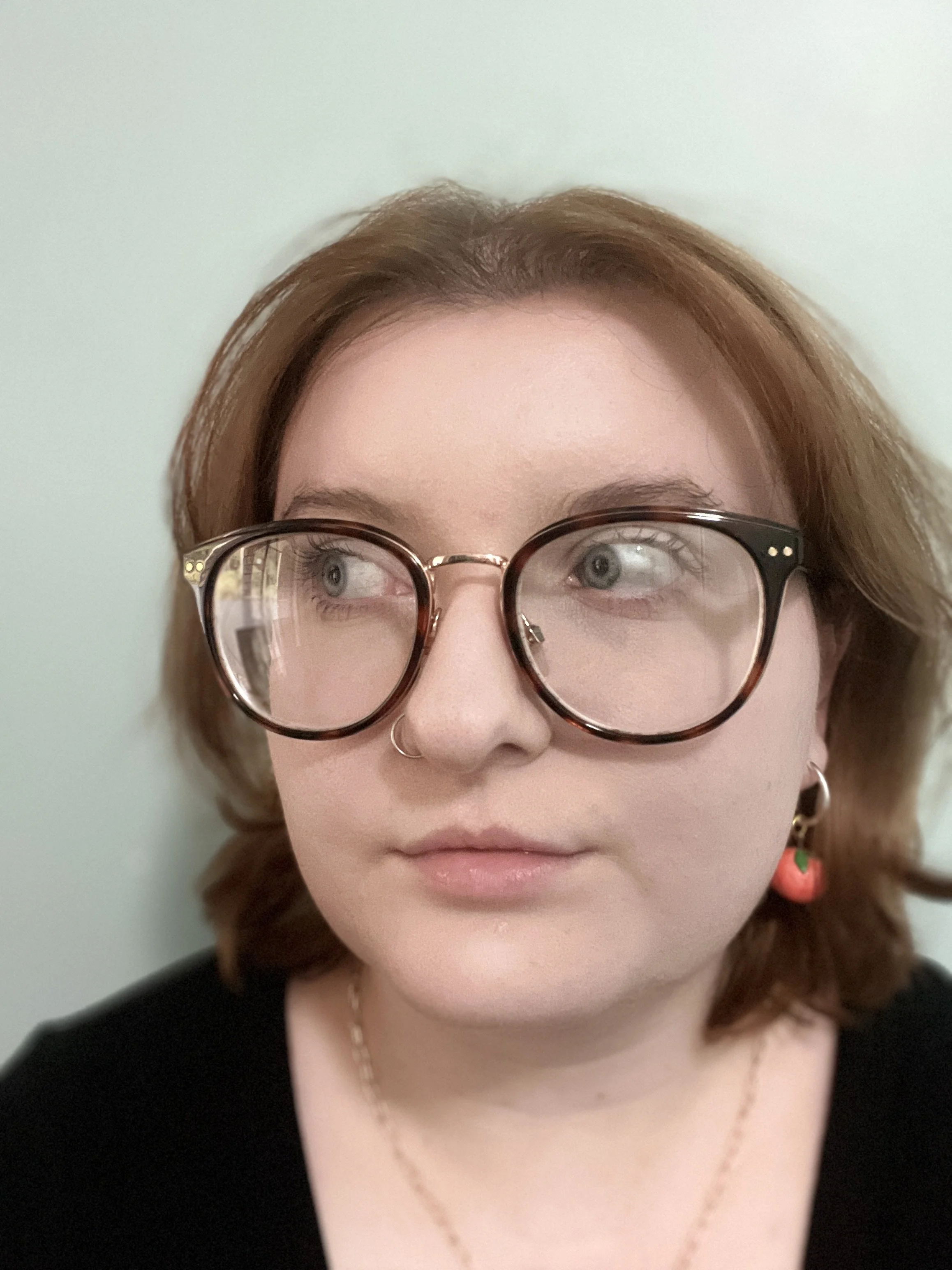 A woman with light skin and auburn hair, wearing large glasses, a nose ring, and strawberry-shaped earrings, looking to her right against a plain pale background.