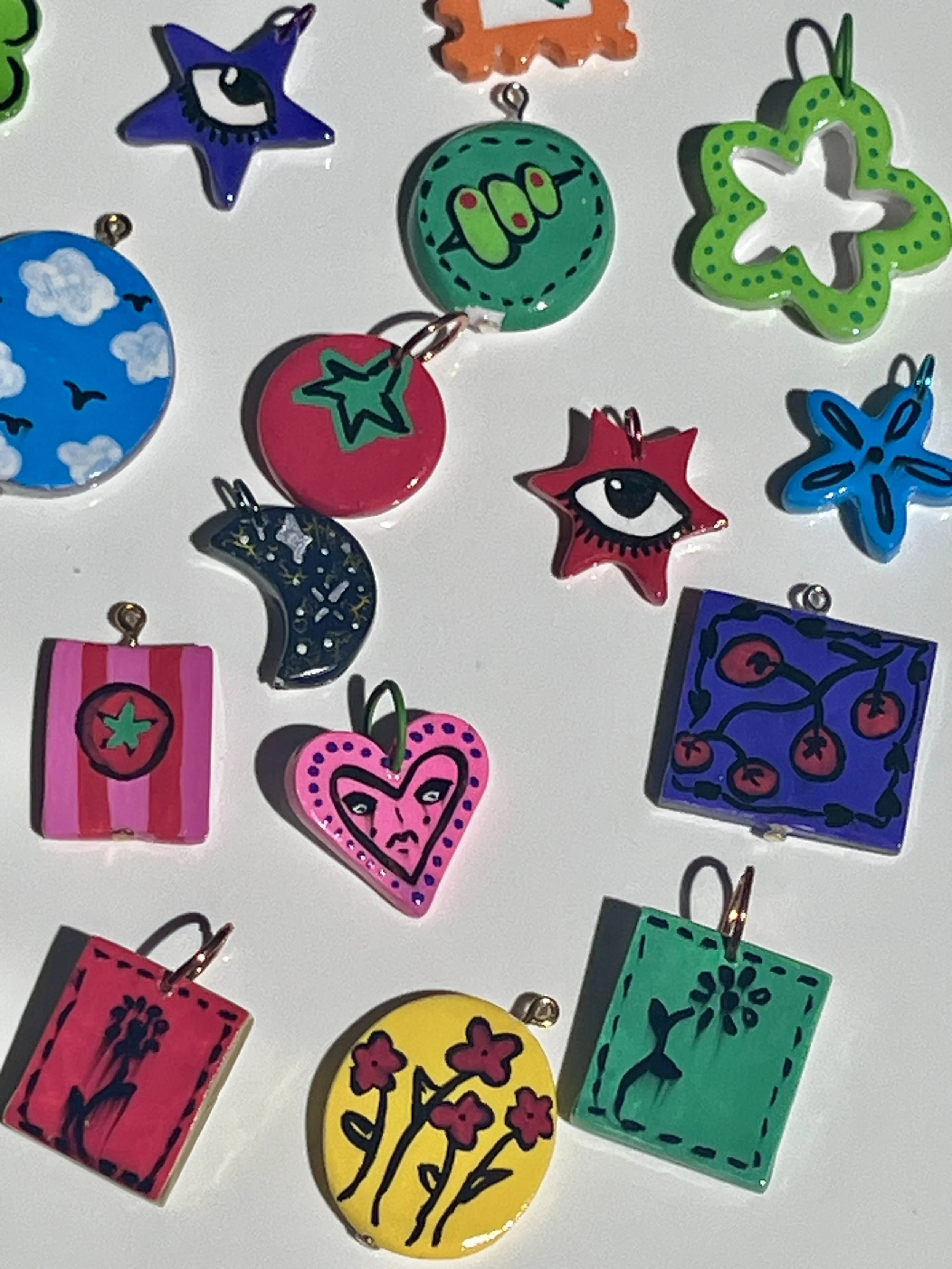 Colorful enamel pins with various shapes and designs, including stars, hearts, moon, flowers, and cherries, arranged on a white surface.