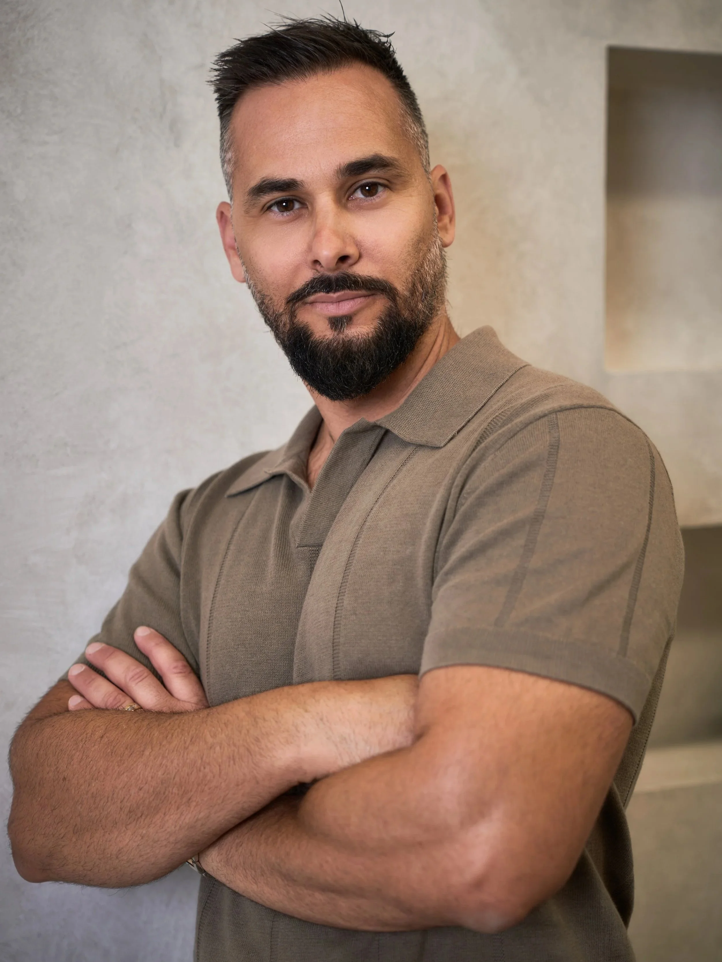 Portrait of a man with a beard crossing his arms, wearing a brown polo shirt, standing against a textured wall.