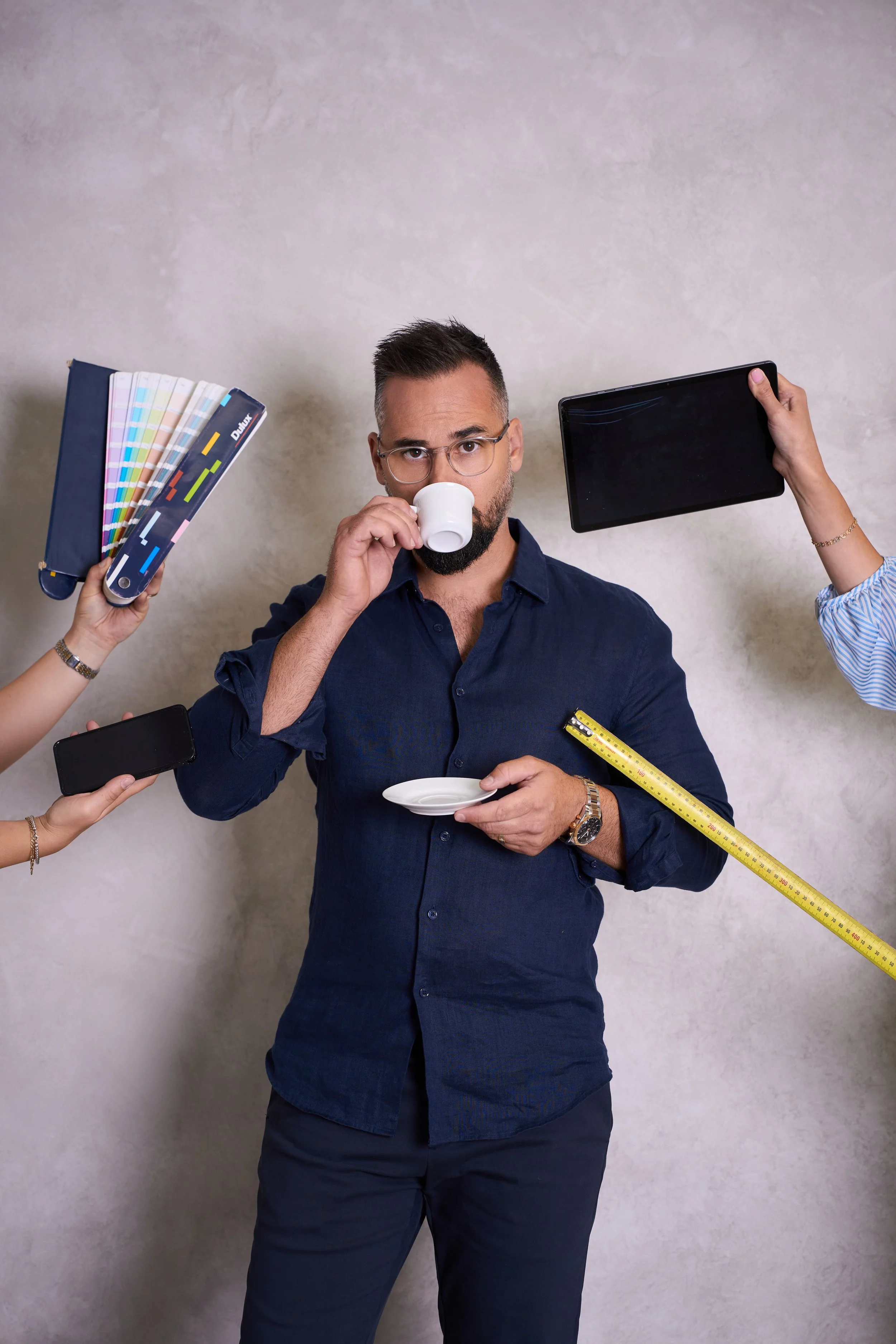 A man in a dark blue shirt standing against a plain wall, drinking from a small cup, surrounded by hands holding color swatch books, a smartphone, a tablet, a measuring tape, and a small plate.