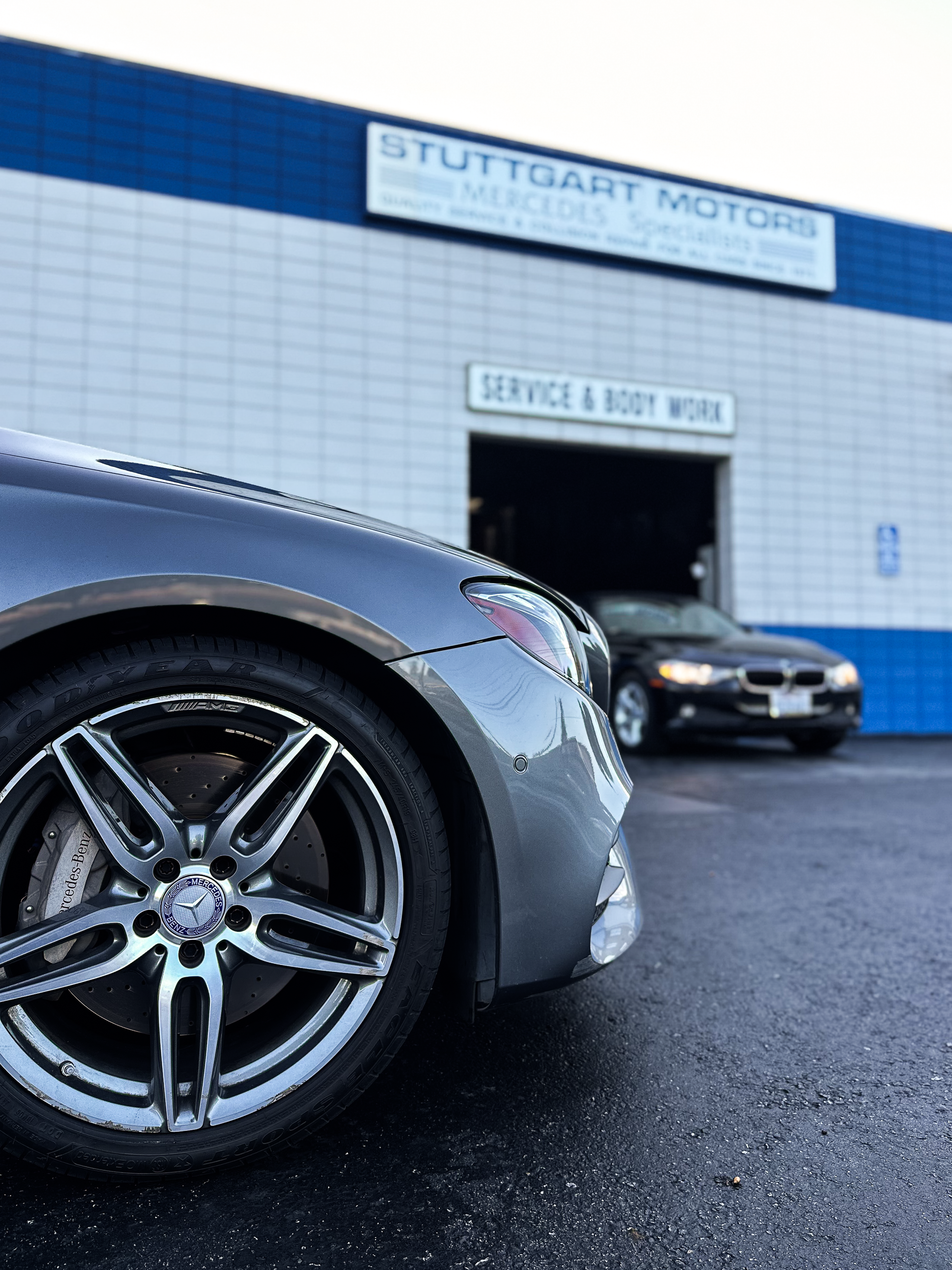 Close-up of a grey Mercedes-Benz car with a focus on the front wheel, parked in front of an automotive service building with a sign reading 'Stuttgart Motors' and 'Service & Body Work' on the exterior.