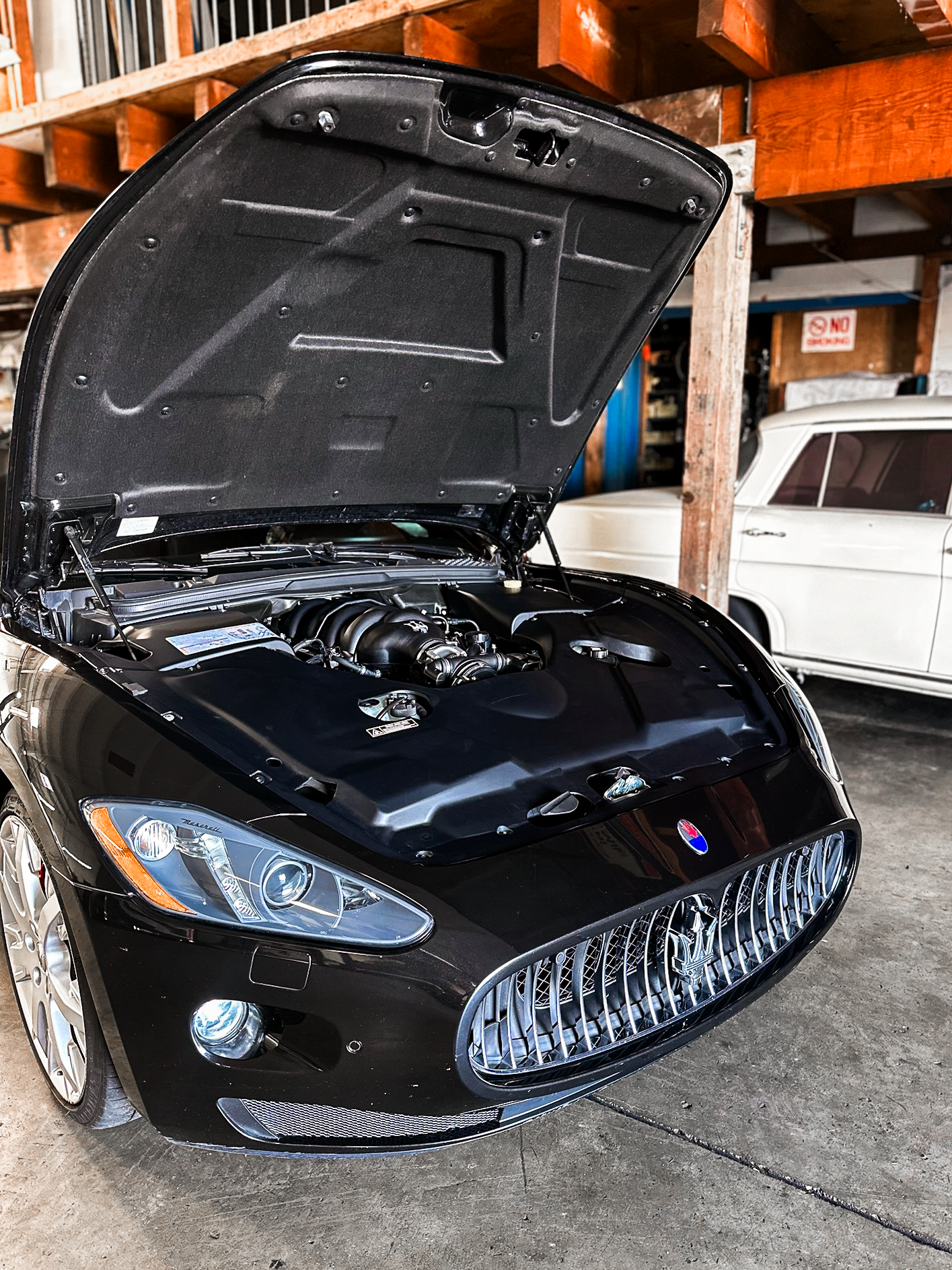 A black Maserati sports car with its hood open, showing the engine. The car is in a garage next to a white vintage car. The Maserati features a prominent front grille and sleek headlights.