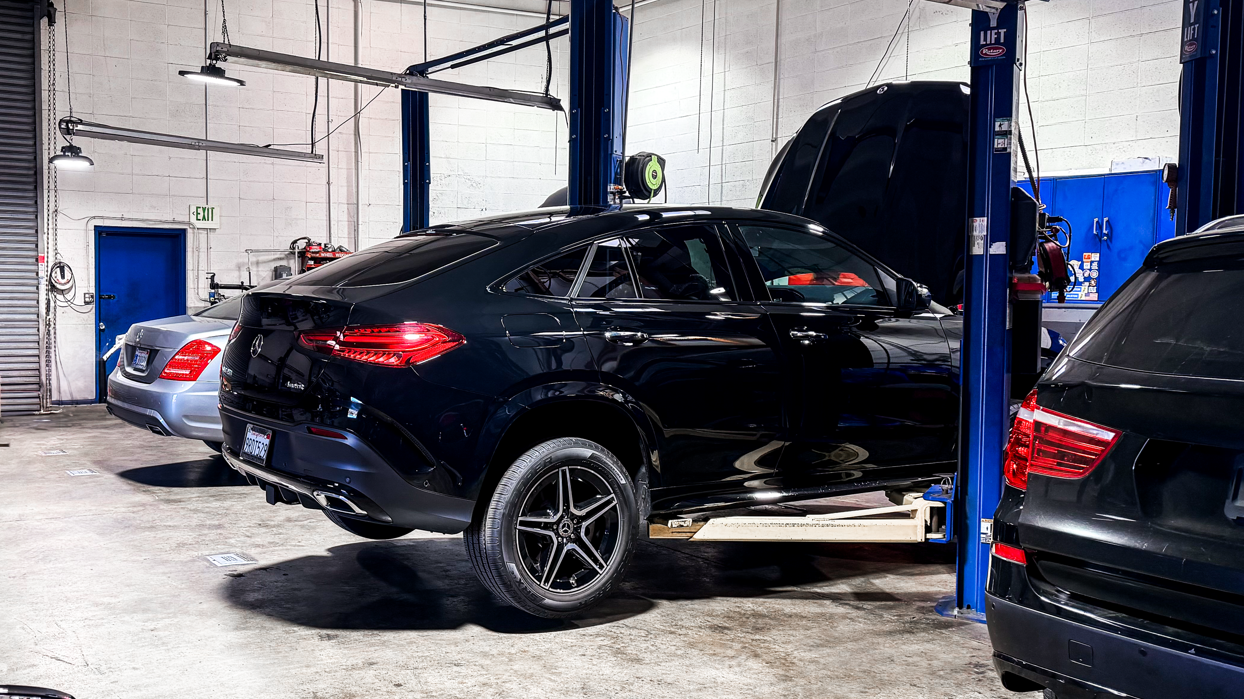 A black Mercedes-Benz car inside an auto repair garage, with its hood open and vehicle lift equipment around.