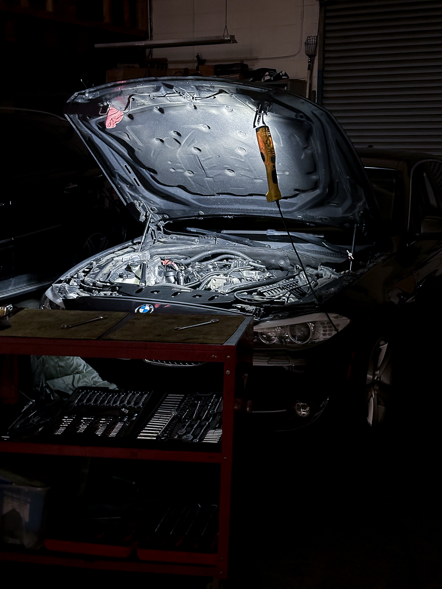 A black BMW with its hood open inside a workshop, revealing the engine, with a yellow tool hanging from the hood. A red workbench with tools is in front of the car and a closed garage door is in the background.