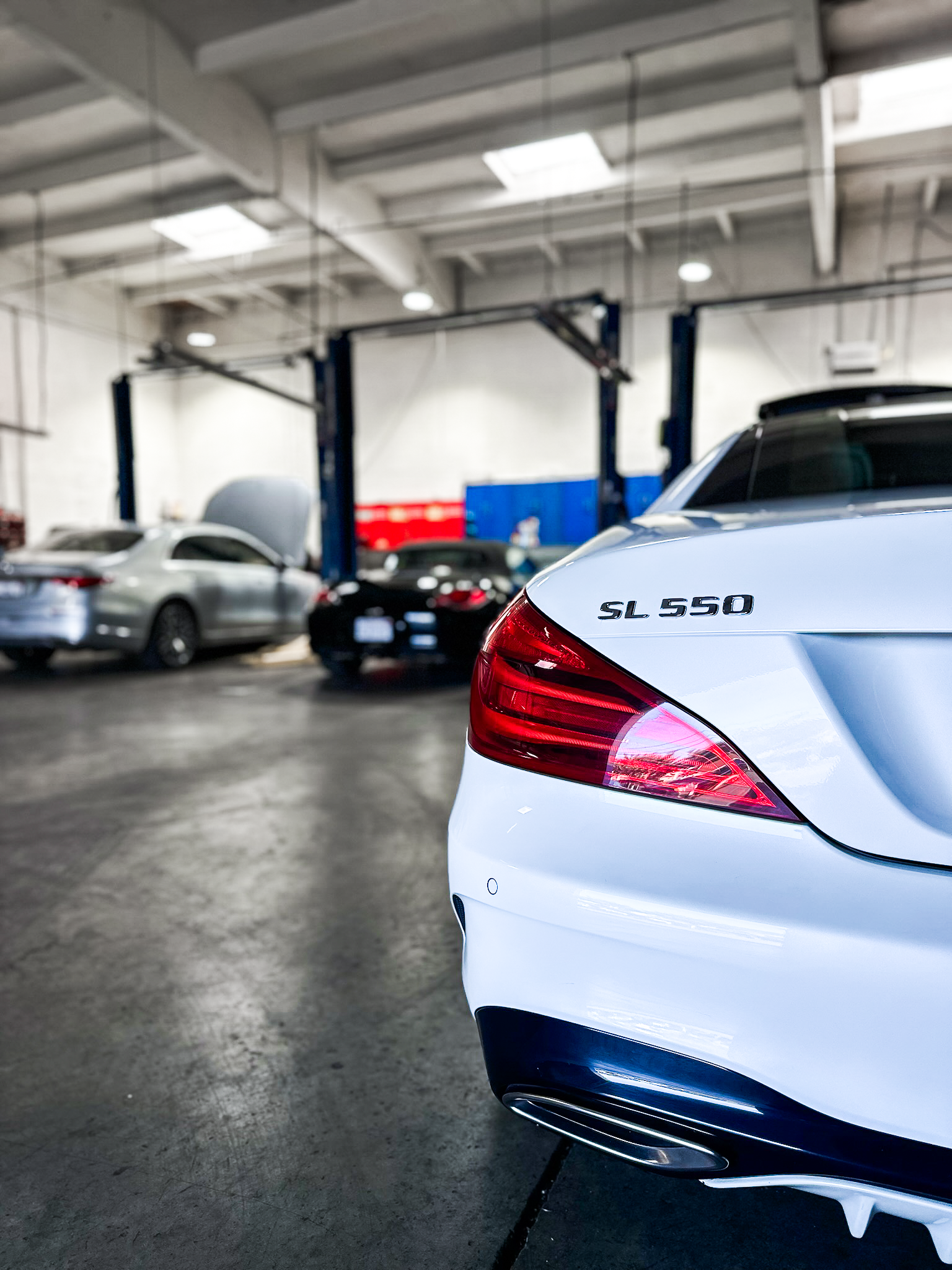 Close-up of a white Mercedes-Benz SL 550 parked inside an automotive service or repair shop, with other cars and equipment visible in the background.