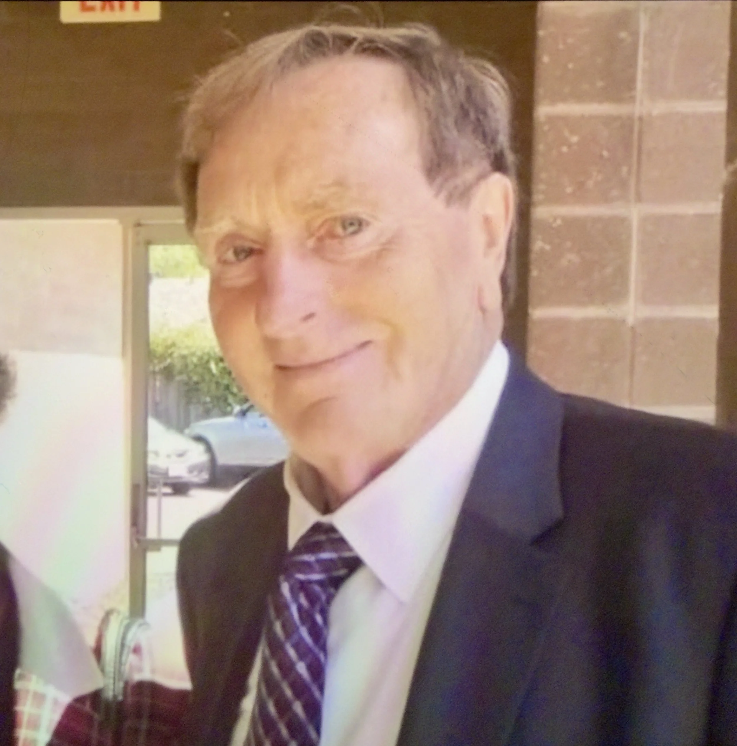 A middle-aged man in formal attire smiling at the camera, wearing a black suit, white shirt, and a purple and white patterned tie, standing indoors with a brick wall and a window in the background.