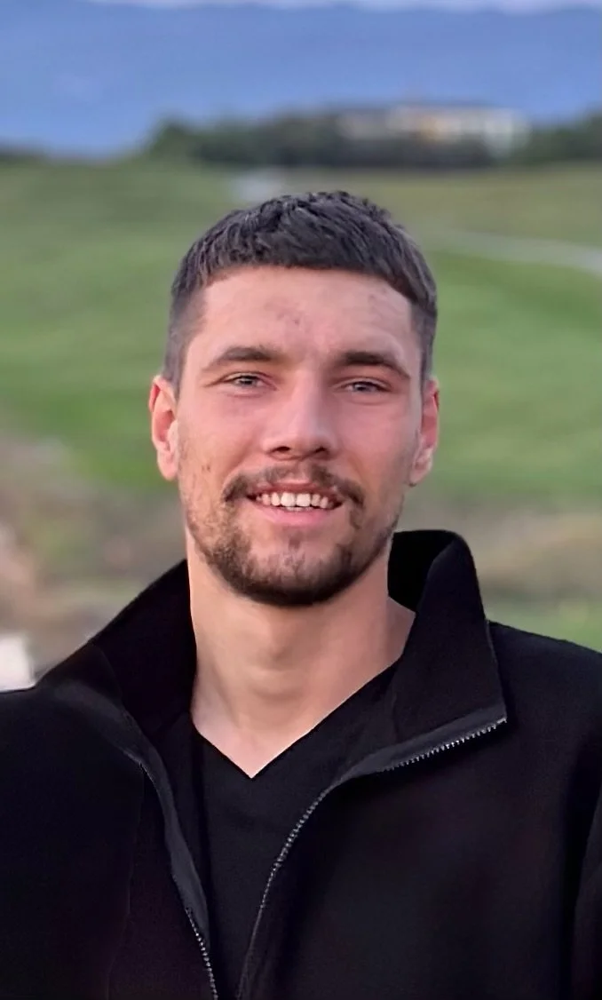 A young man with short dark hair and a beard, smiling outdoors with a blurred green landscape and blue sky in the background.