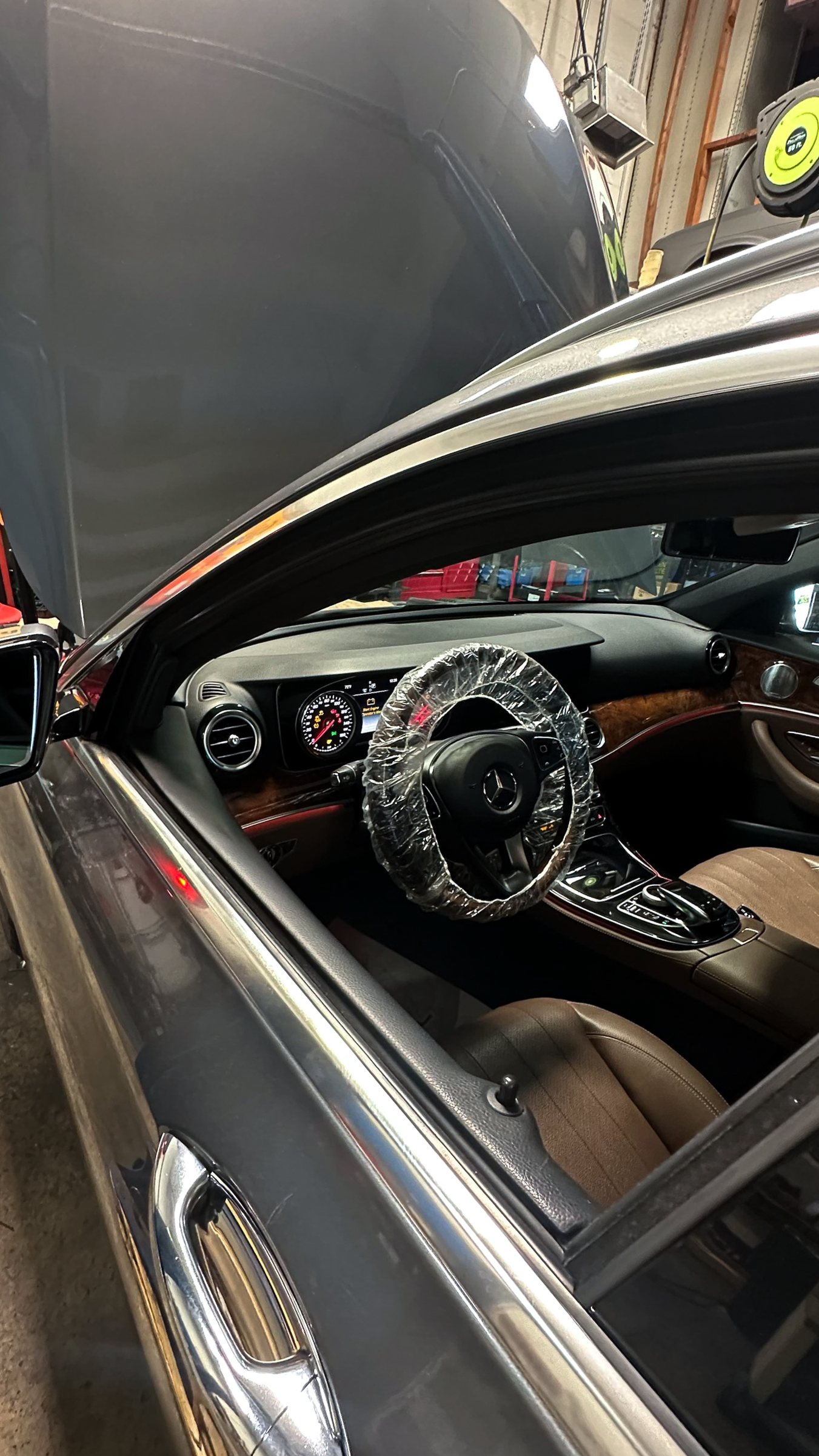 Inside a gray Mercedes-Benz car with a plastic-covered steering wheel, displayed in a garage with open hood and various tools visible in the background.