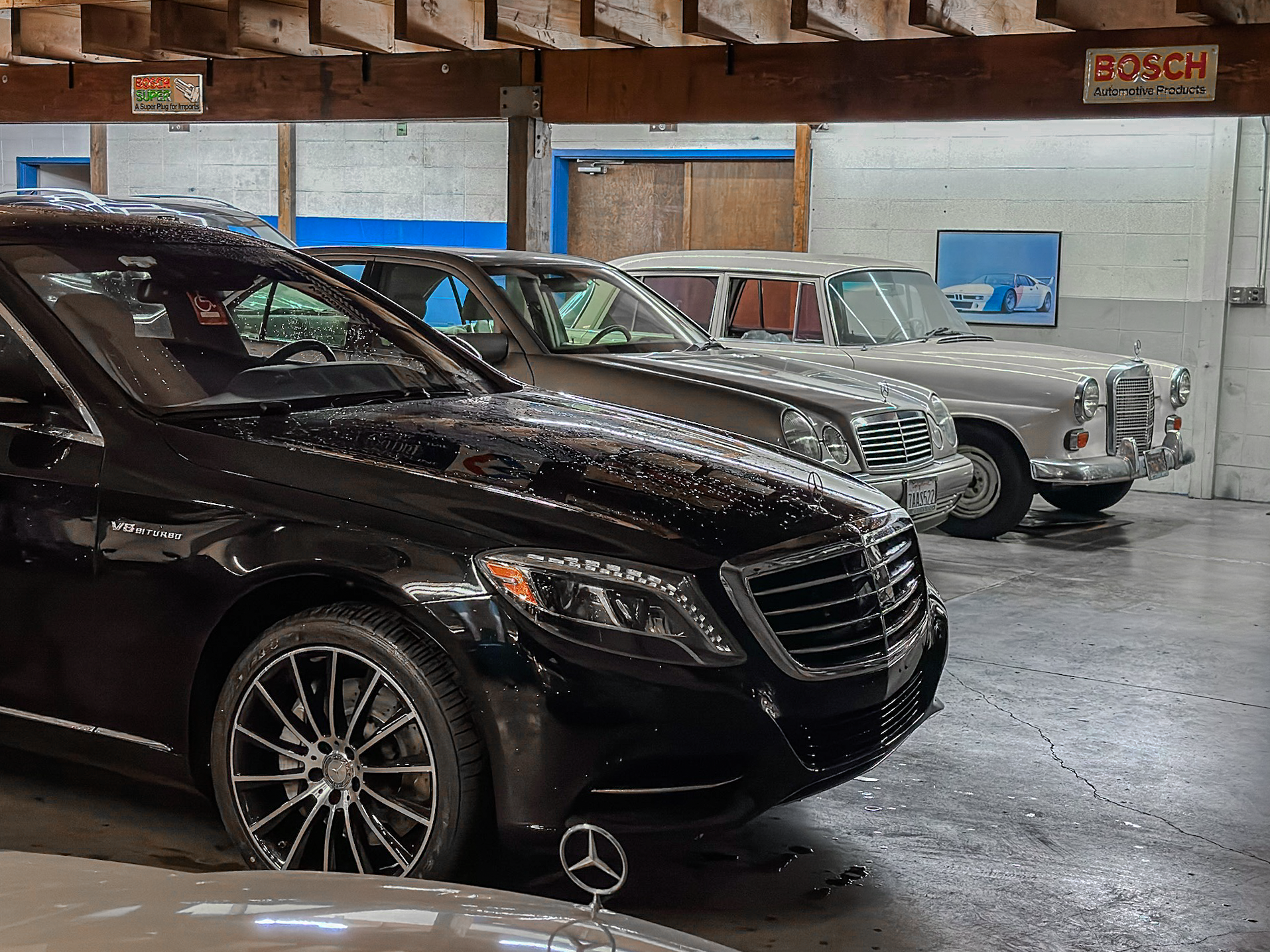 Interior of a garage with four classic and luxury cars parked side by side, including a black Mercedes-Benz with AMG branding, a gray Mercedes-Benz sedan, and a vintage silver Mercedes sedan. The garage has signs from Bosch and other automotive brand
