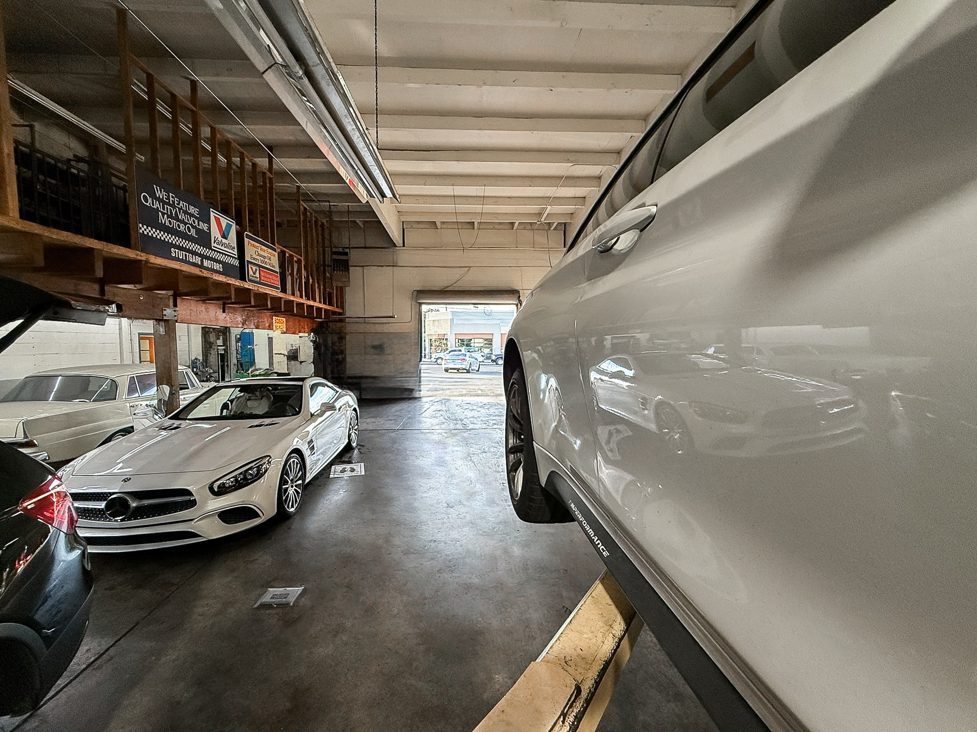 View of an indoor parking garage with several classic and luxury cars, including a white Mercedes-Benz and a white SUV, with some cars parked outside visible through the open garage door.