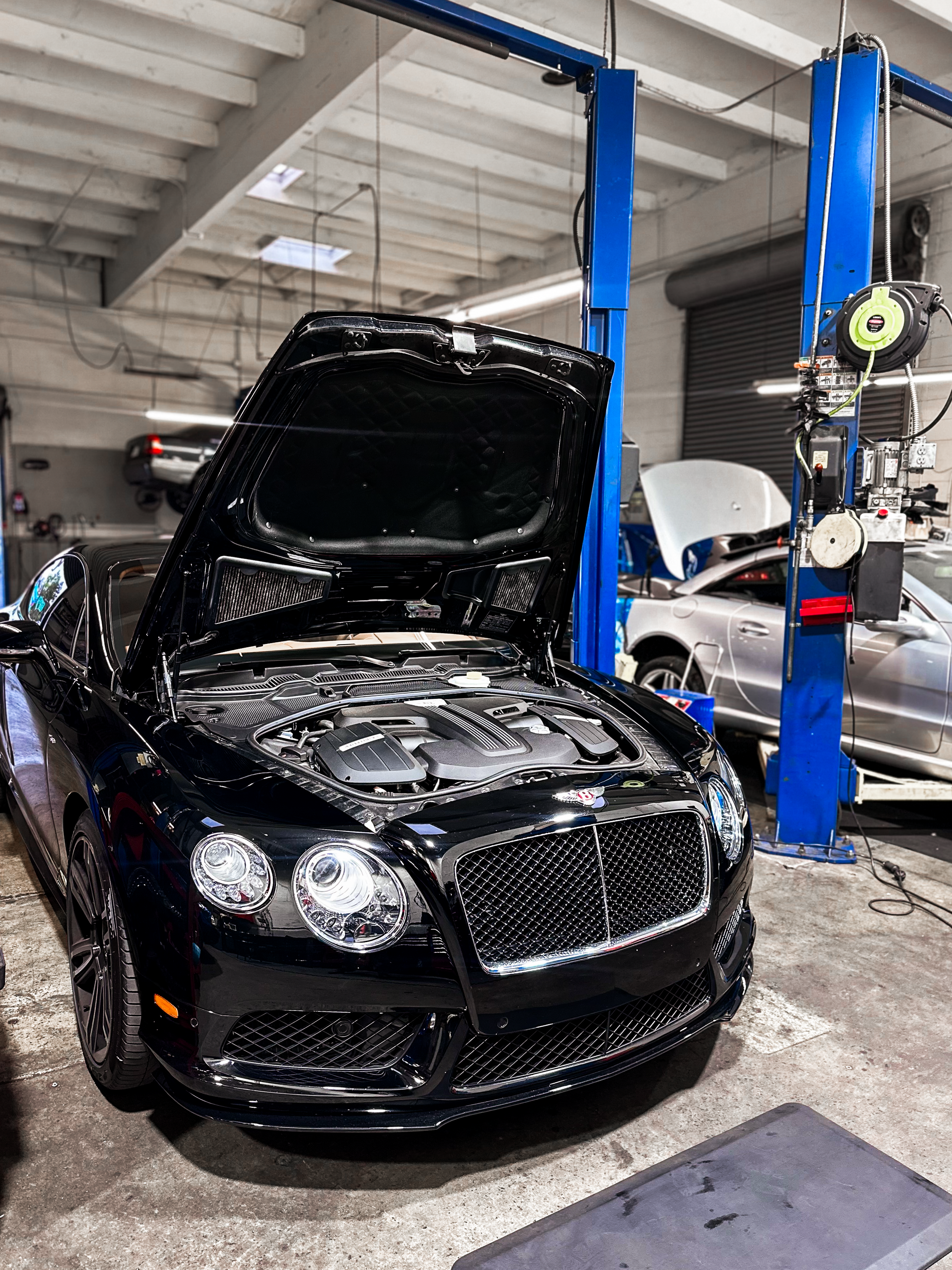 Black luxury car with open hood in an auto repair shop, surrounded by repair equipment and other vehicles.