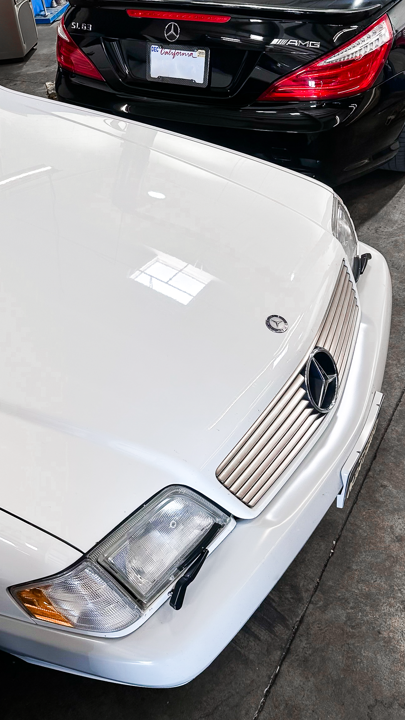 Close-up of the front of a white vintage Mercedes-Benz car with a distinctive grille and emblem, parked indoors next to a black Mercedes-Benz car with an California license plate.