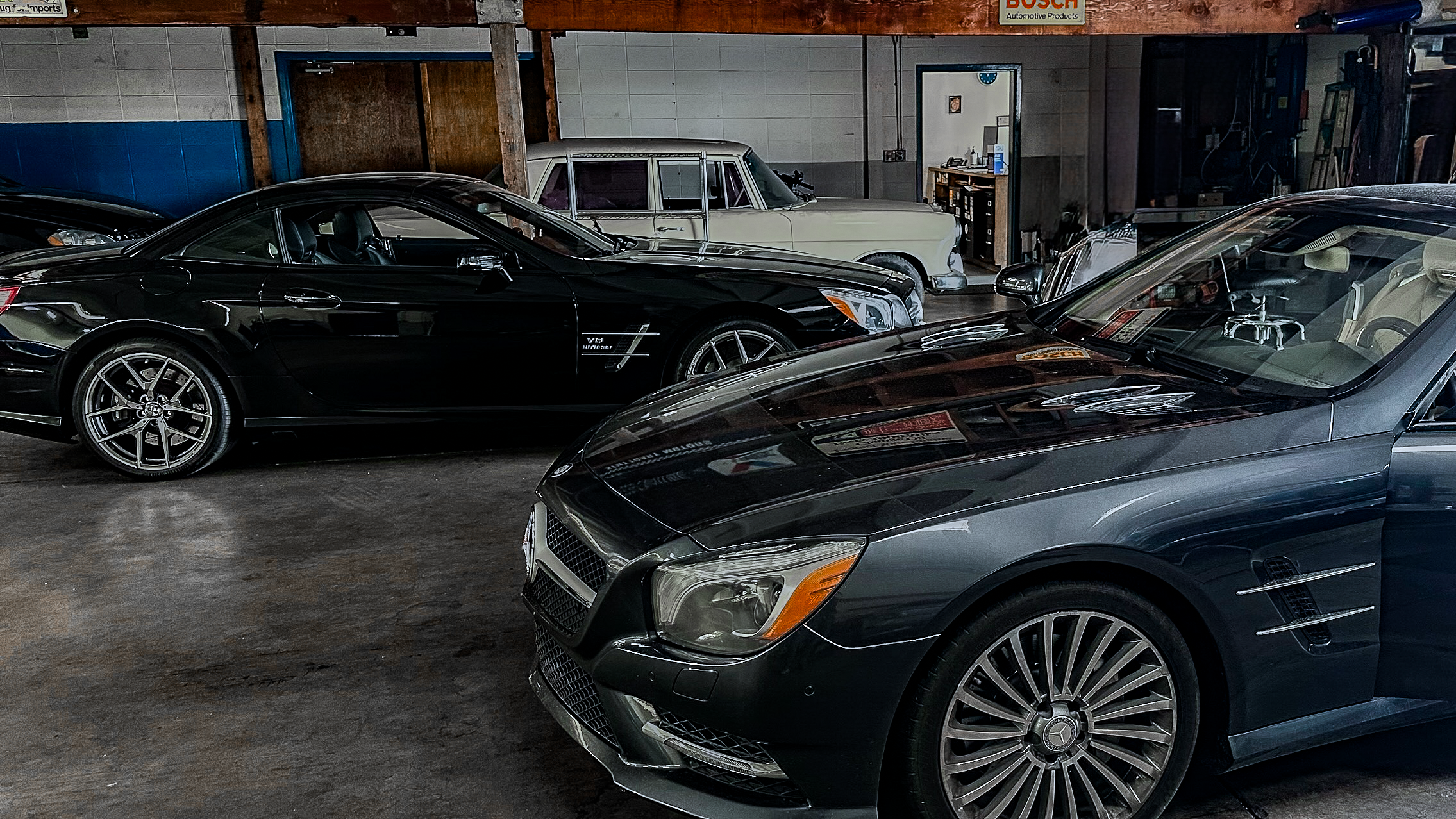 Multiple cars parked inside a garage, including a black sports car and a gray luxury car in the foreground, with an older white car in the background.