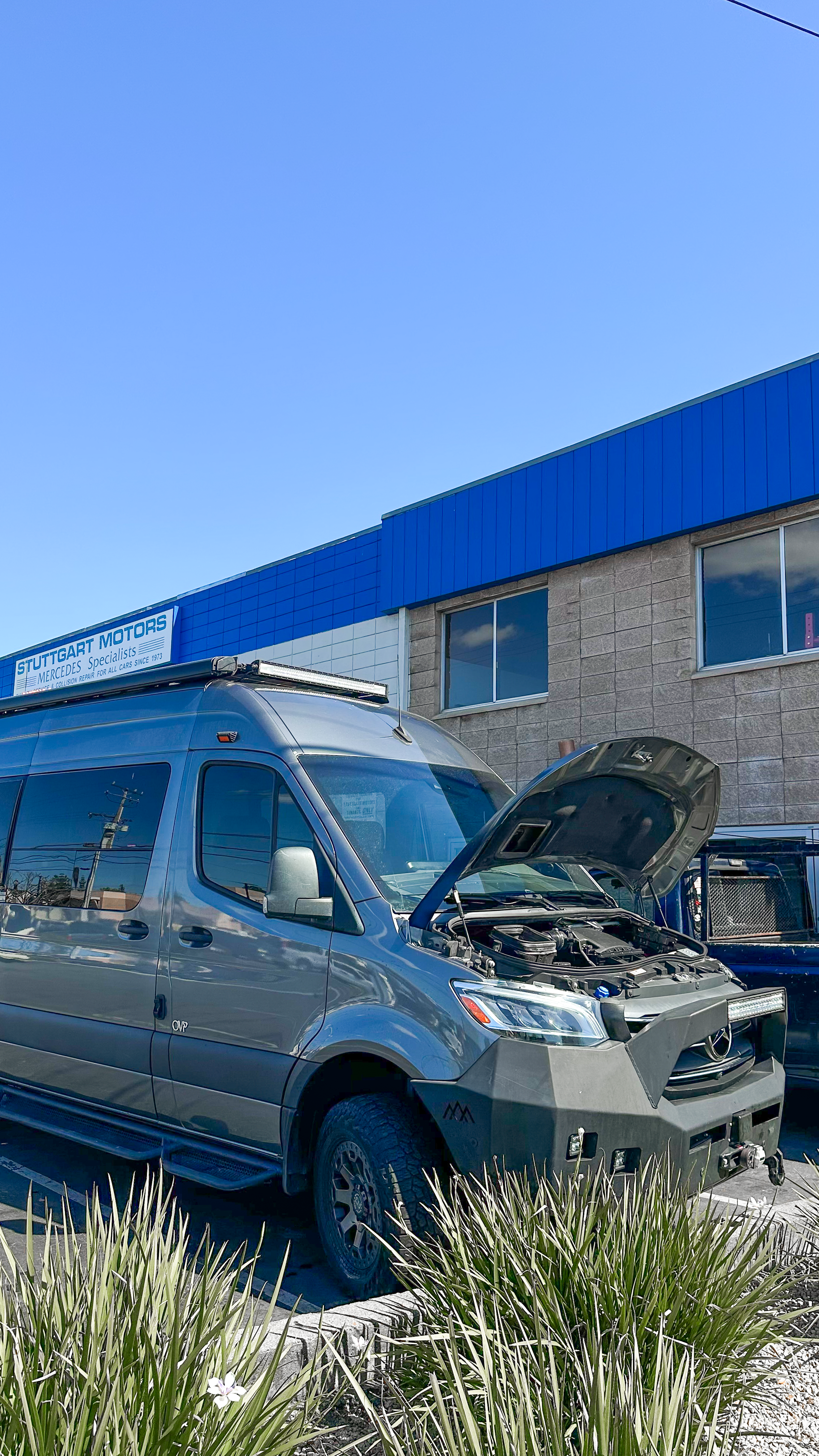 Silver van with hood open parked outside a building with a blue and gray exterior sign that reads 'Stuttgart Motors Mercedes Specialist' under a clear blue sky.