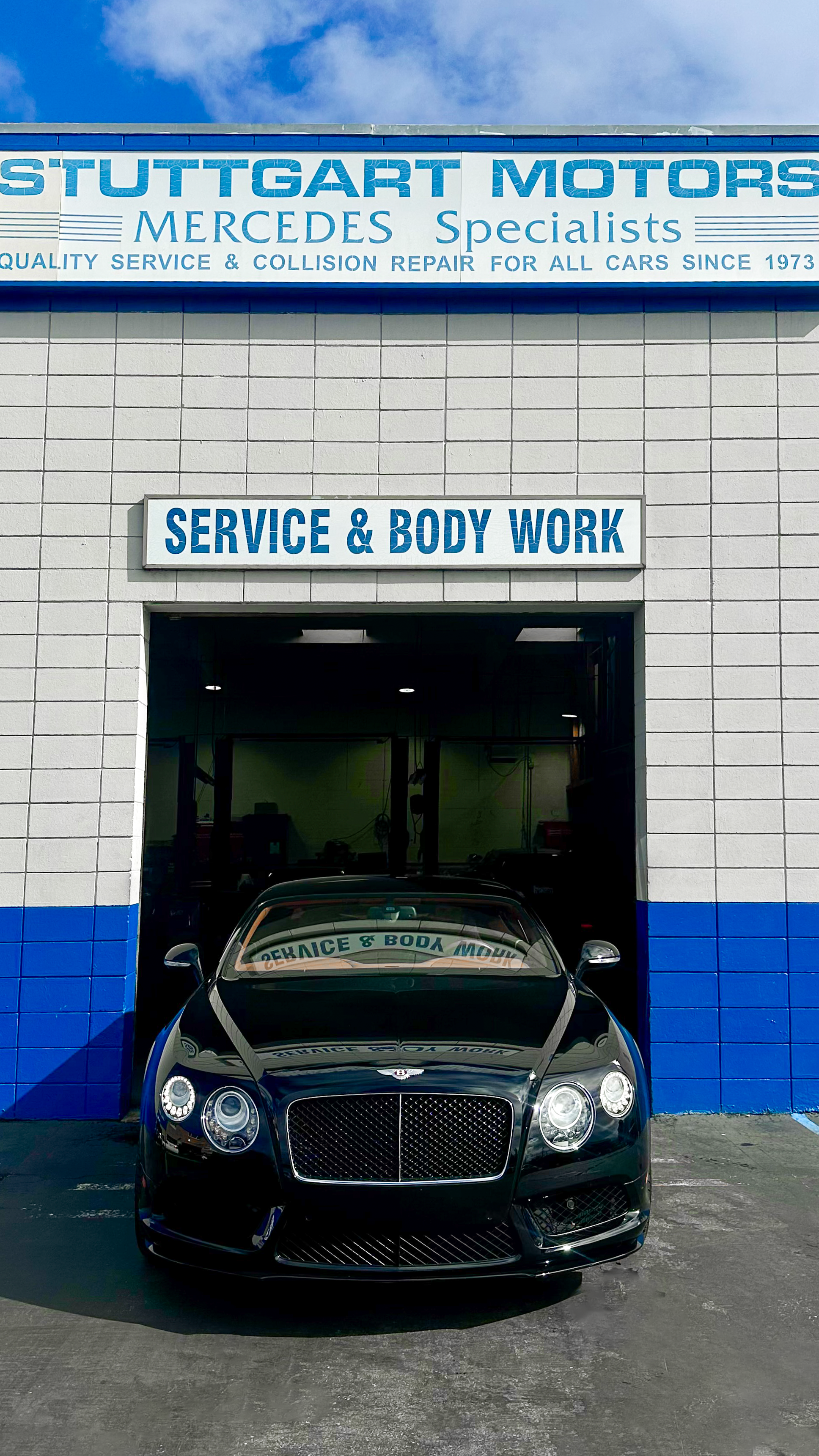 Black luxury car parked in front of an auto repair shop with signs indicating service and body work, and specialization in Mercedes vehicles.