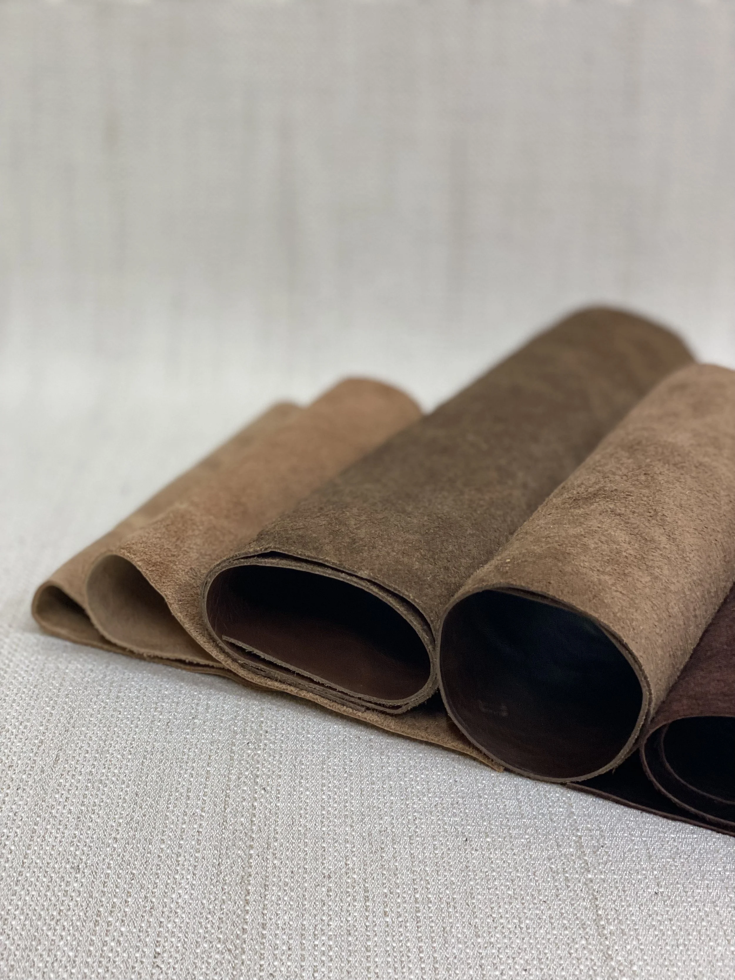 Three rolled pieces of leather or suede material in shades of brown, placed on a light-colored fabric surface.