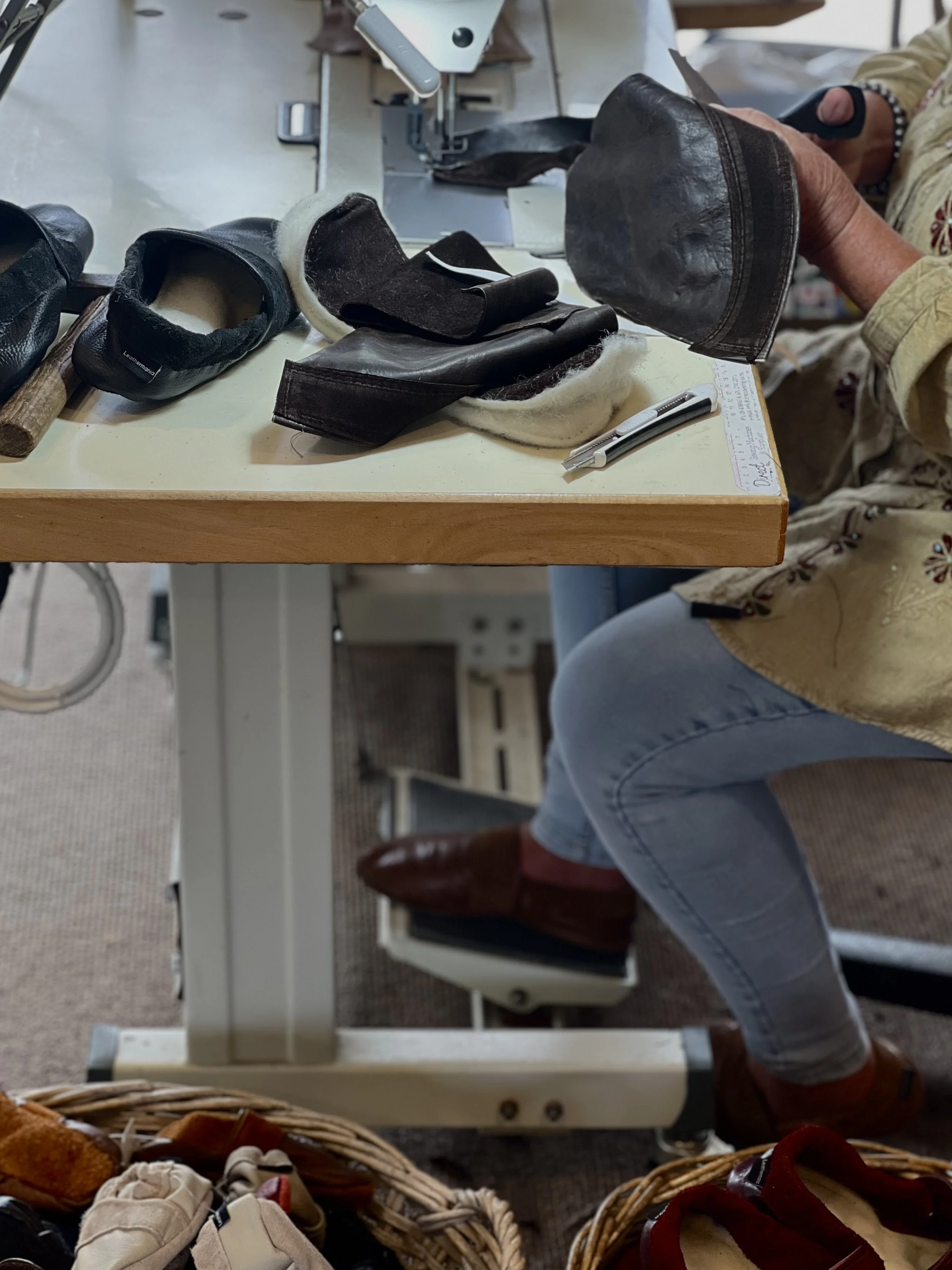 A person working on shoe repair or leather craft at a workstation with various leather shoes, tools, and materials on the table and in baskets.