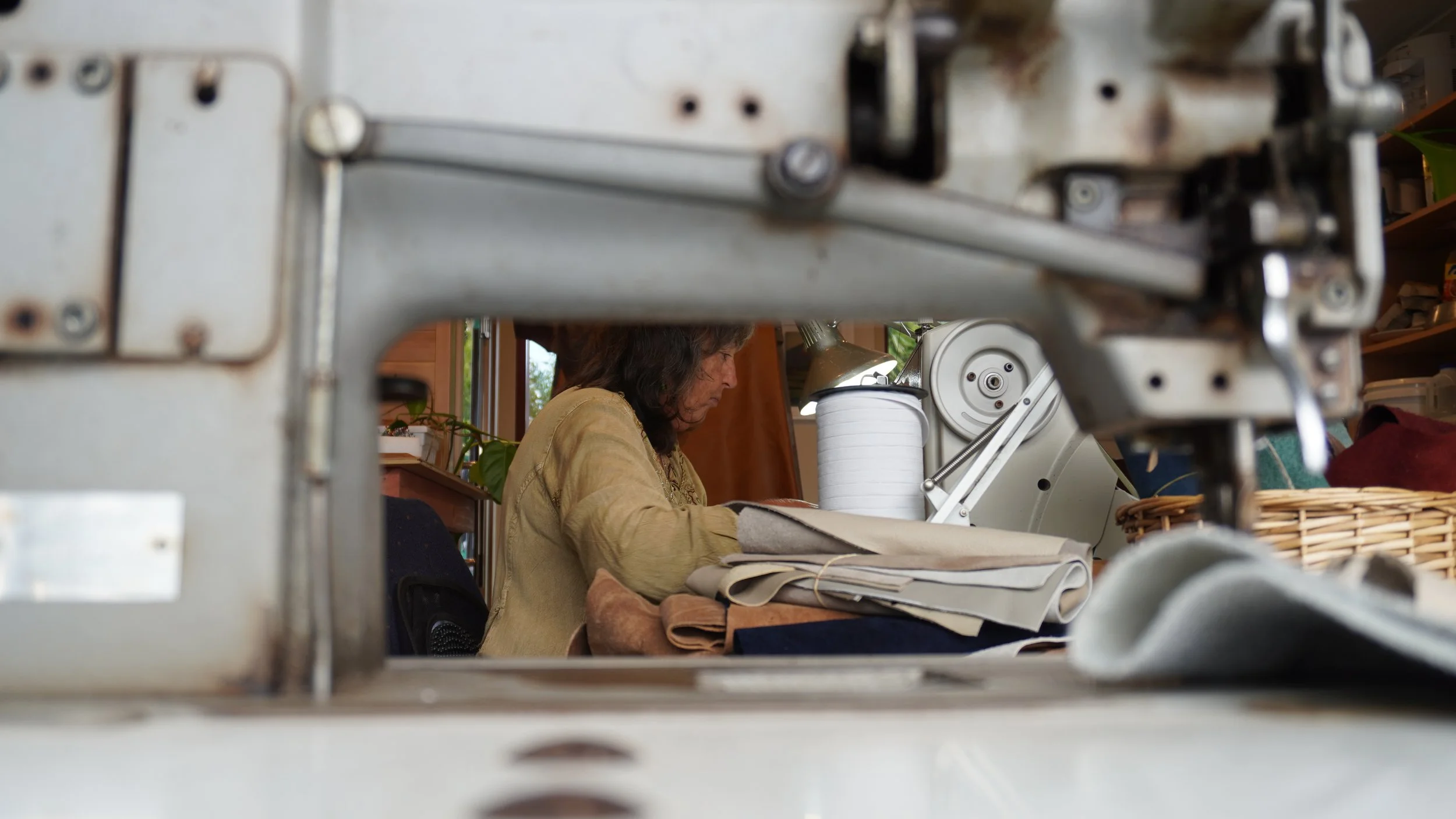 A woman working at a desk, viewed through a metal machine or device with bolts and screws in the foreground, surrounded by sewing or crafting materials.