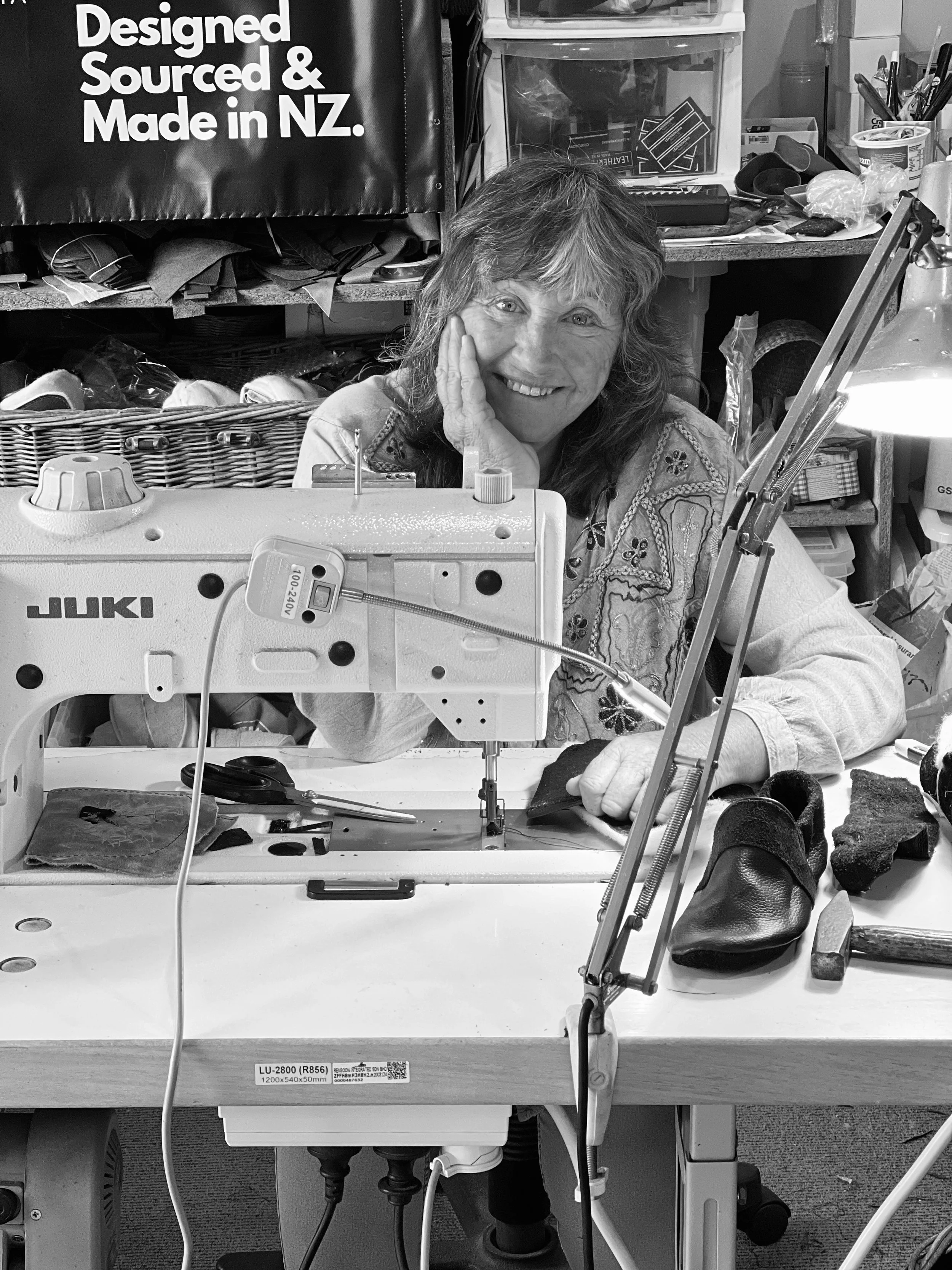 A woman smiling and resting her face on her hand while working at a sewing machine in a cluttered workshop. The workspace contains various tools, fabrics, and supplies, with a storage bin and a sign that reads 'Designed, Sourced & Made in NZ.'