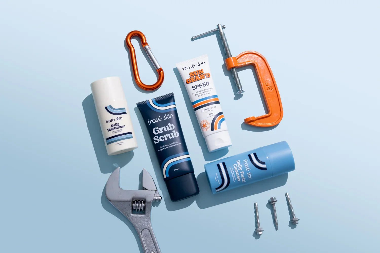 Assorted skincare products, tools, and screws arranged on a light blue surface. Items include a white bottle labeled 'Daily Moisturiser', a navy tube labeled 'Grub Scrub', a white tube labeled 'Sun Guard SPF 50', a blue bottle labeled 'Daily Face & Body Scrub', an orange carabiner, an orange clamp, a small silver clamp, an adjustable wrench, an Allen wrench, and three screws.